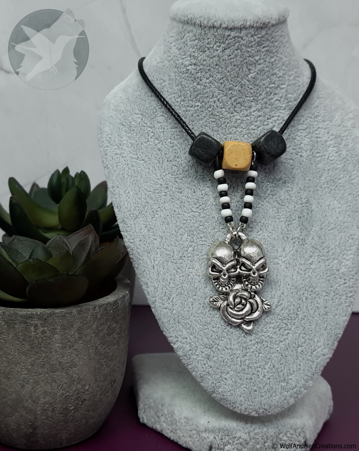 Necklace with skull and rose pendant on a gray stand next to a potted plant.
