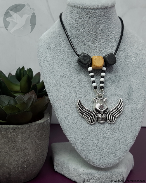 Necklace with skull and wings pendant on a gray stand next to a potted plant.