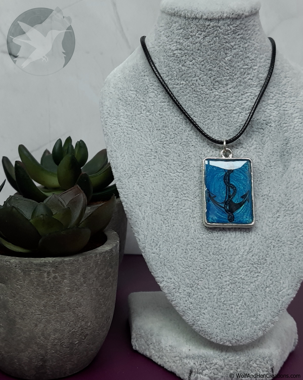 A rectangular pendant necklace displayed on a grey fabric mannequin bust, featuring a blue and black anchor design on a black cord. A small potted succulent sits to the left.