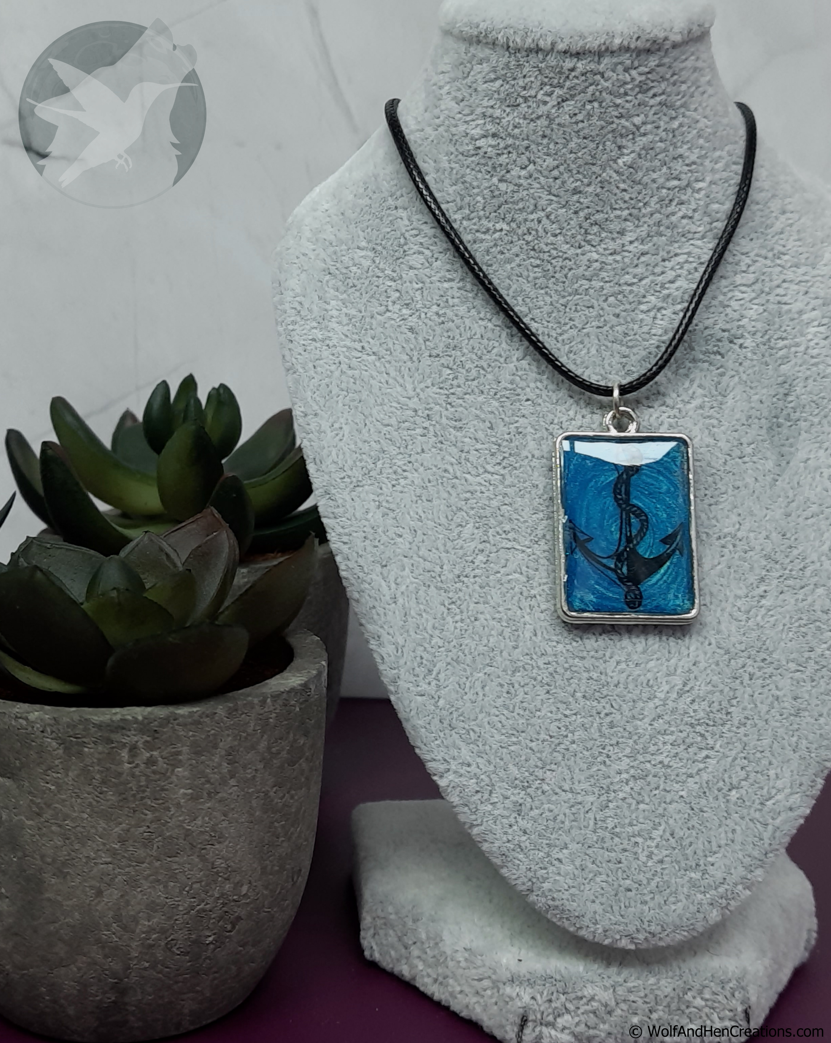 A rectangular pendant necklace displayed on a grey fabric mannequin bust, featuring a blue and black anchor design on a black cord. A small potted succulent sits to the left.