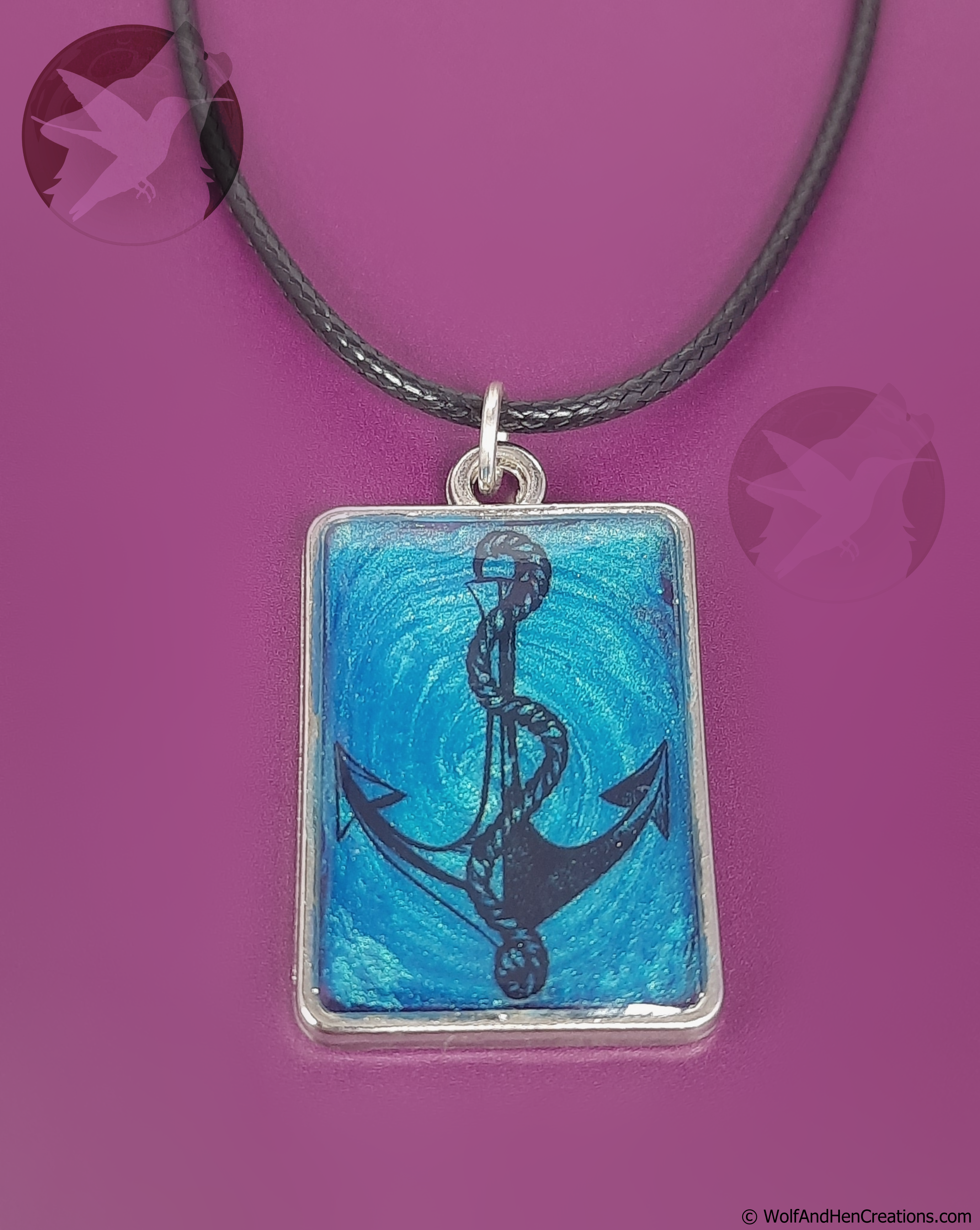 A rectangular pendant necklace featuring a black anchor illustration over a textured blue background, suspended from a black cord on a solid magenta background.