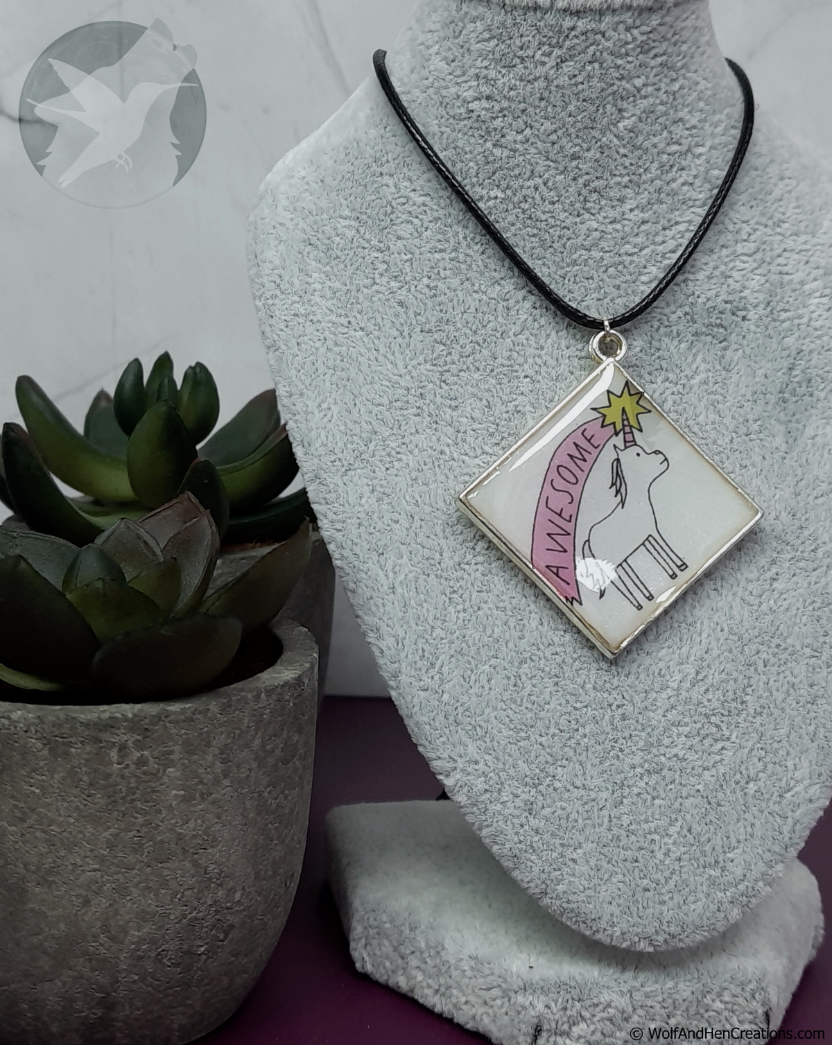 A square pendant necklace displayed on a grey fabric mannequin bust, featuring a black cord and a white pendant with a silver-toned border. The pendant has a black line drawing of a unicorn looking up at a yellow shooting star over it's horn, with the word 'AWESOME' in the pink banner-like trail