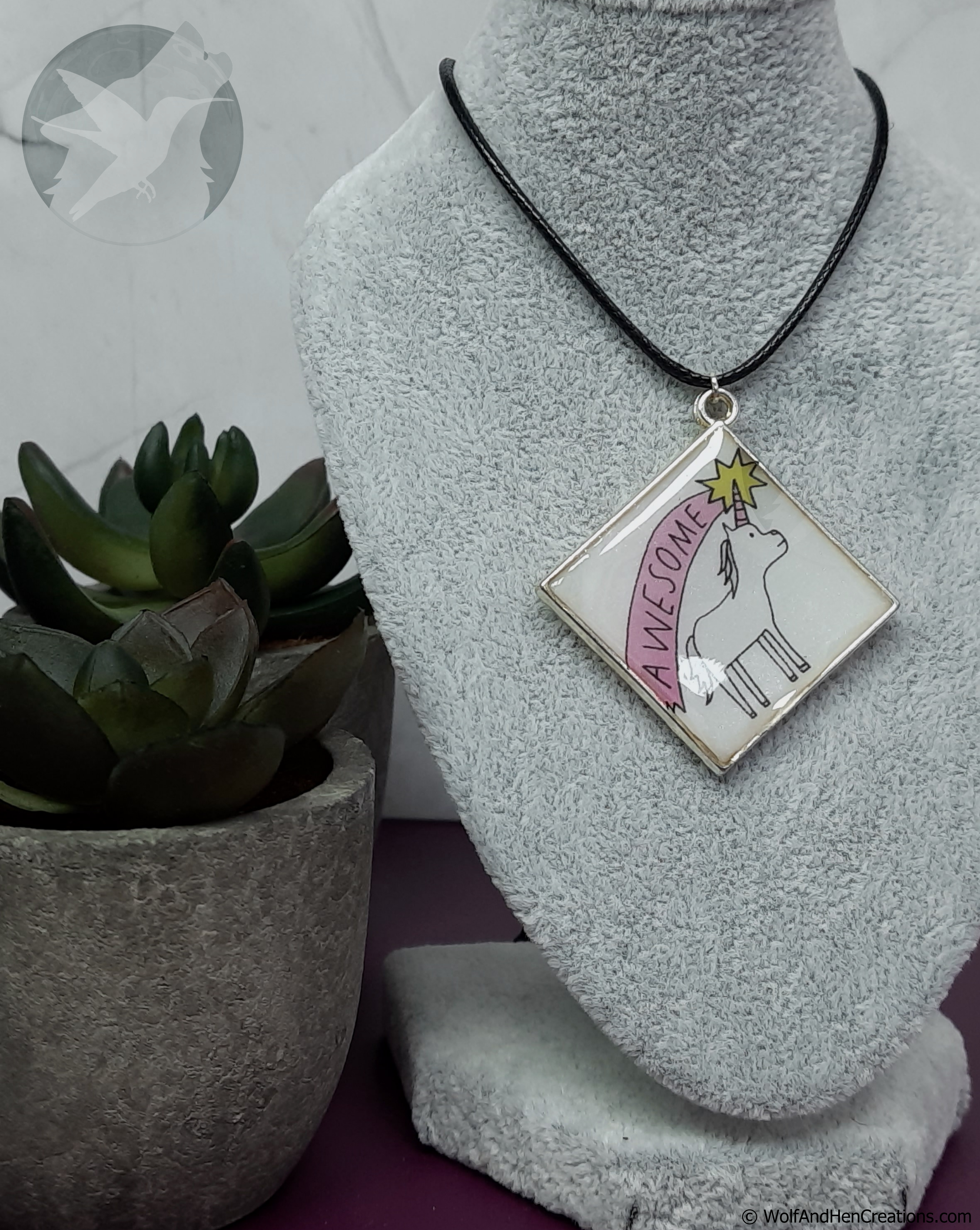 A square pendant necklace displayed on a grey fabric mannequin bust, featuring a black cord and a white pendant with a silver-toned border. The pendant has a black line drawing of a unicorn looking up at a yellow shooting star over it's horn, with the word 'AWESOME' in the pink banner-like trail
