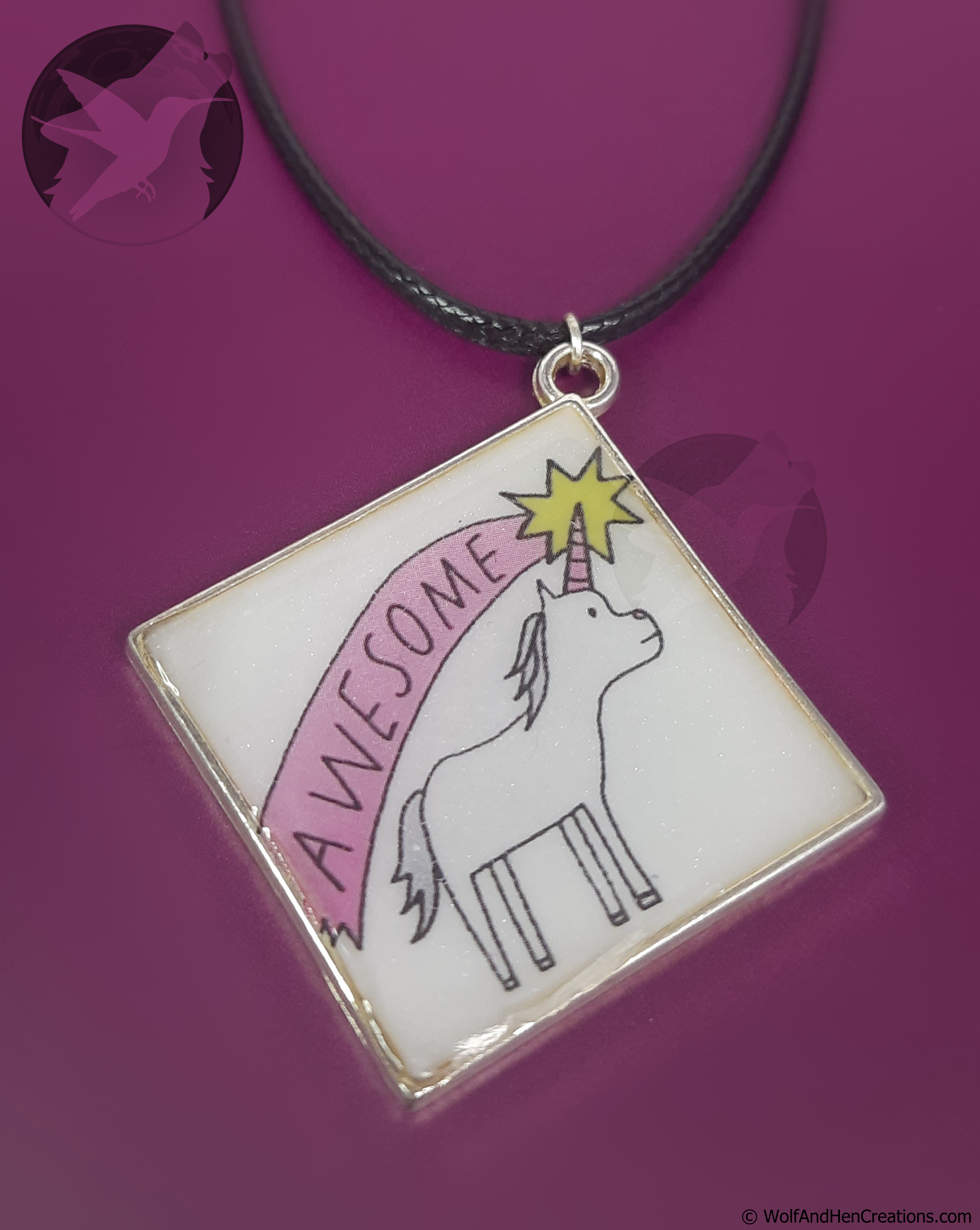 A square pendant featuring a white unicorn with a pink horn under a pink arch and yellow starburst that says AWESOME, hanging from a black cord against a purple background.