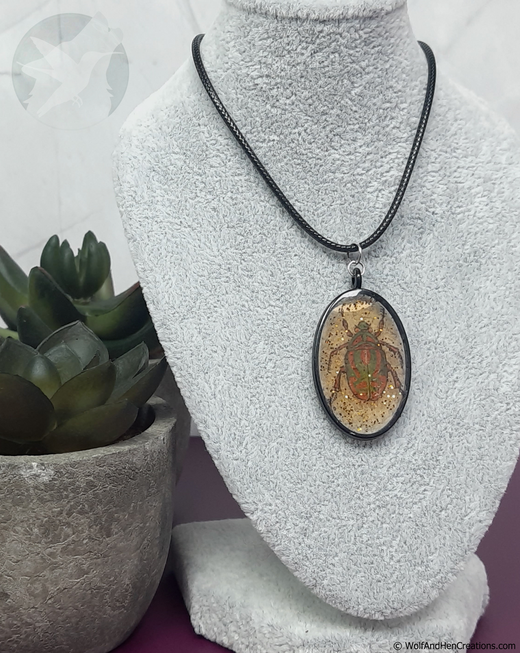 A black cord necklace with an oval pendant featuring a red and green beetle design on a translucent amber-yellow background, displayed on a grey jewellery bust next to a potted succulent.