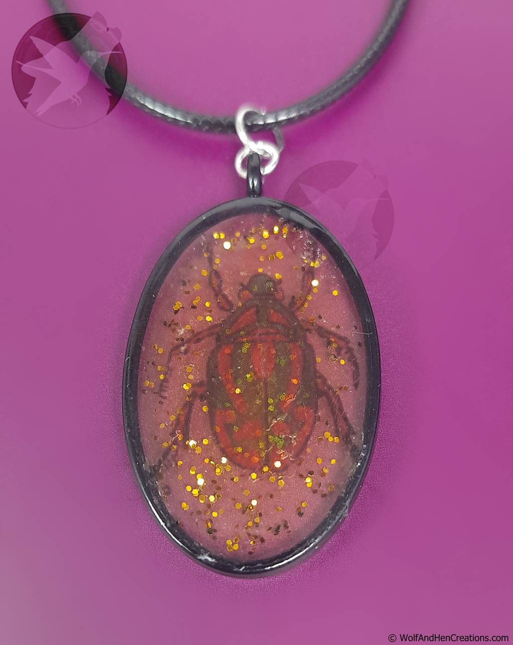 An oval pendant necklace featuring a red and green beetle design inside translucent amber-yellow resin with gold glitter, suspended from a black cord against a solid purple background.