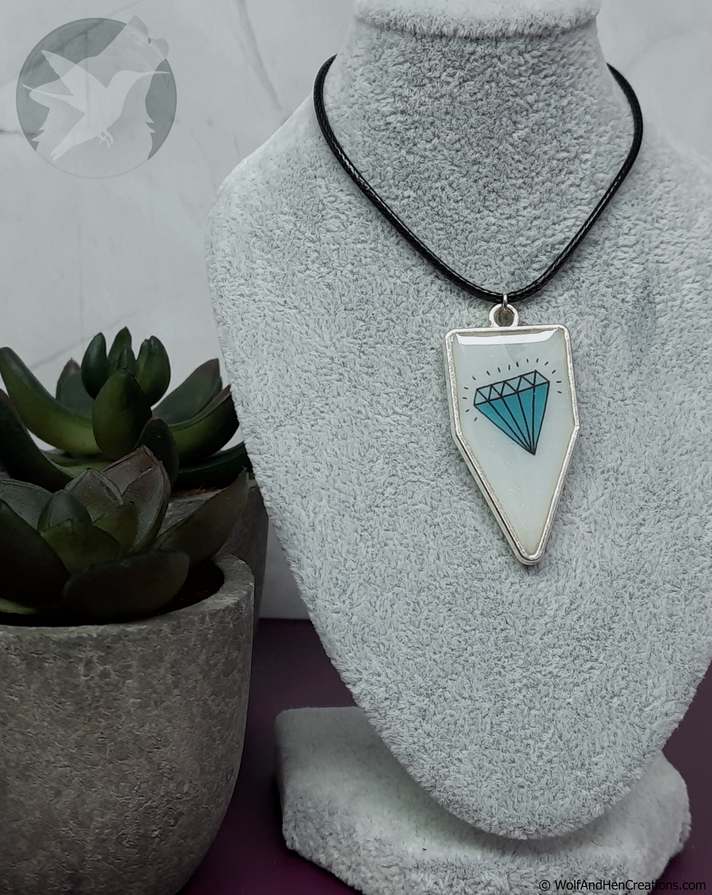 A silver pennant or banner shaped pendant necklace with a white background and a blue diamond graphic design, displayed on a grey velvet mannequin bust next to a potted succulent.