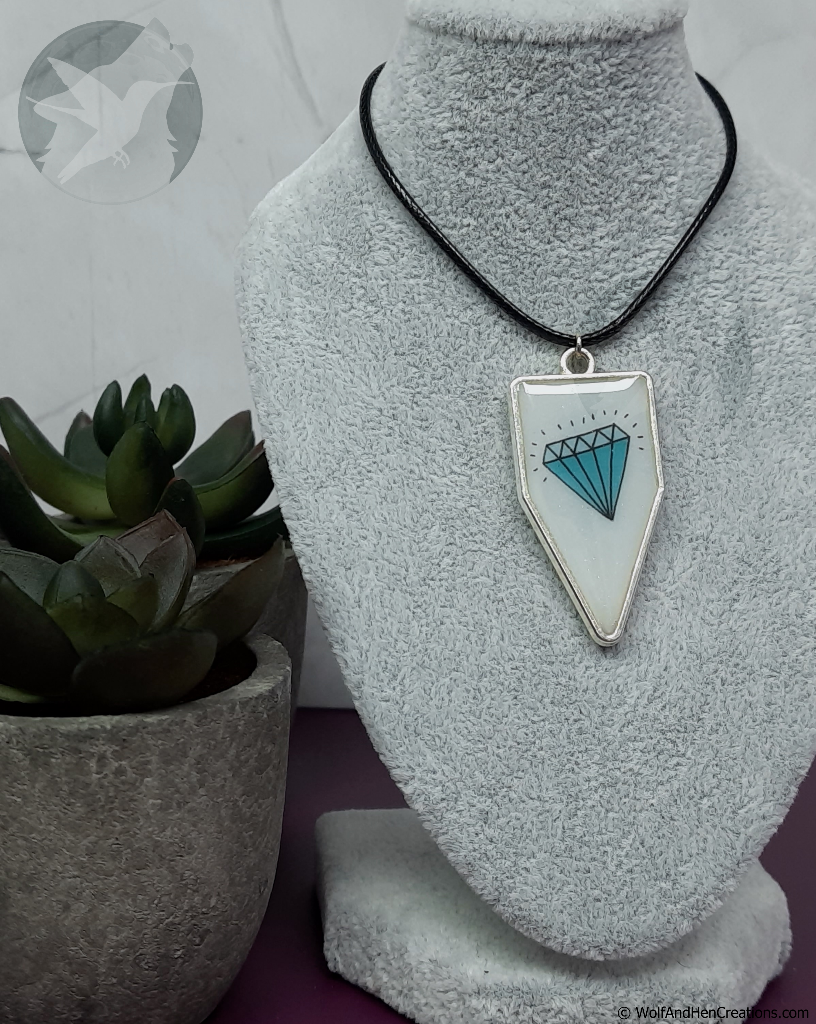 A silver pennant or banner shaped pendant necklace with a white background and a blue diamond graphic design, displayed on a grey velvet mannequin bust next to a potted succulent.