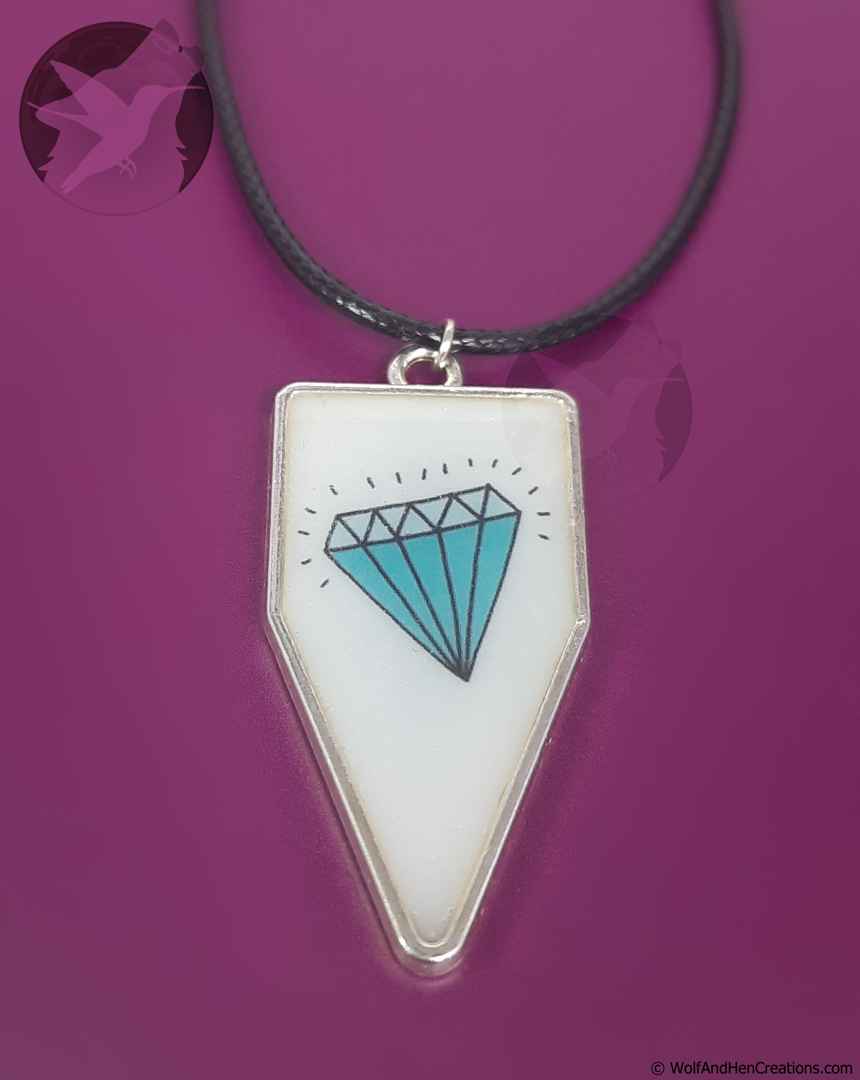 A silver pendant necklace with a pearlescent white pennant or banner shaped face featuring a blue and black diamond illustration, suspended on a black cord against a magenta background.