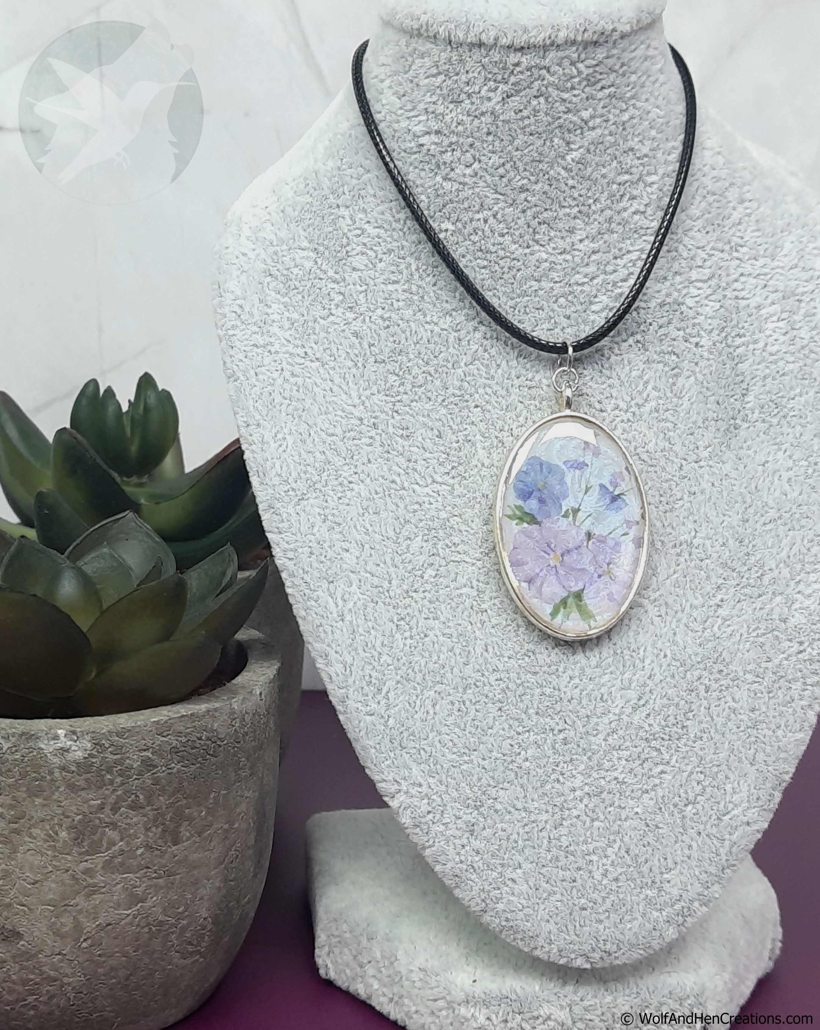 An oval pendant necklace with a silver toned frame and a watercolour style floral design of blue and purple flowers, displayed on a white textured jewellery bust. The necklace is on a black cord, with a potted succulent plant on the left side.