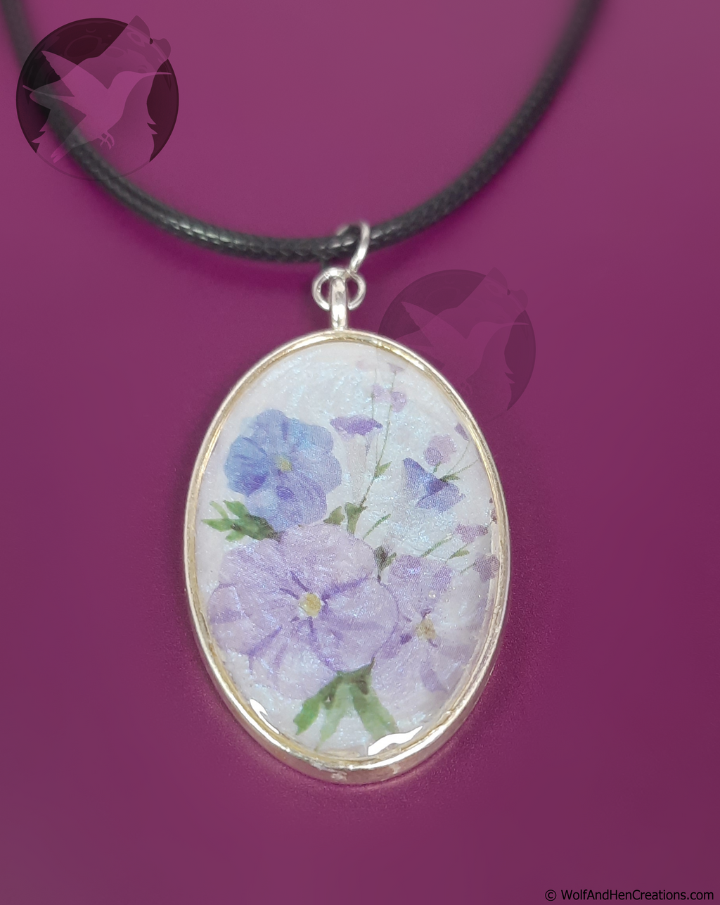 An oval pendant necklace featuring a delicate watercolour floral design of light purple and blue flowers with green leaves, set in a silver toned bezel on a black cord, presented against a solid magenta background.
