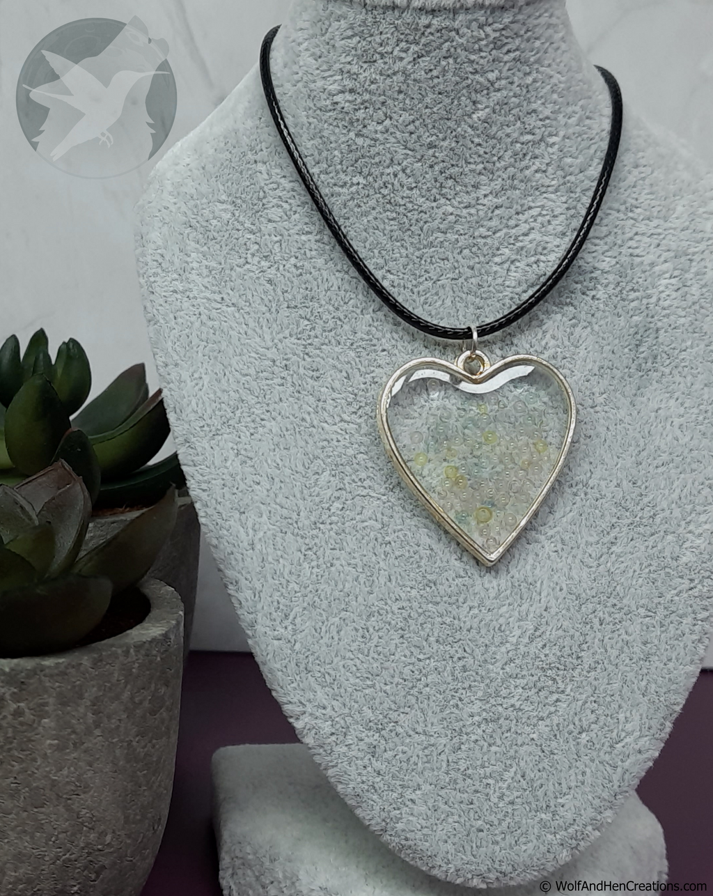 A heart-shaped pendant necklace with a silver-toned metal border and a clear centre filled with multicoloured translucent bubbles. Strung on a black cord, it is displayed on a grey textured jewellery bust with a potted succulent in the background.