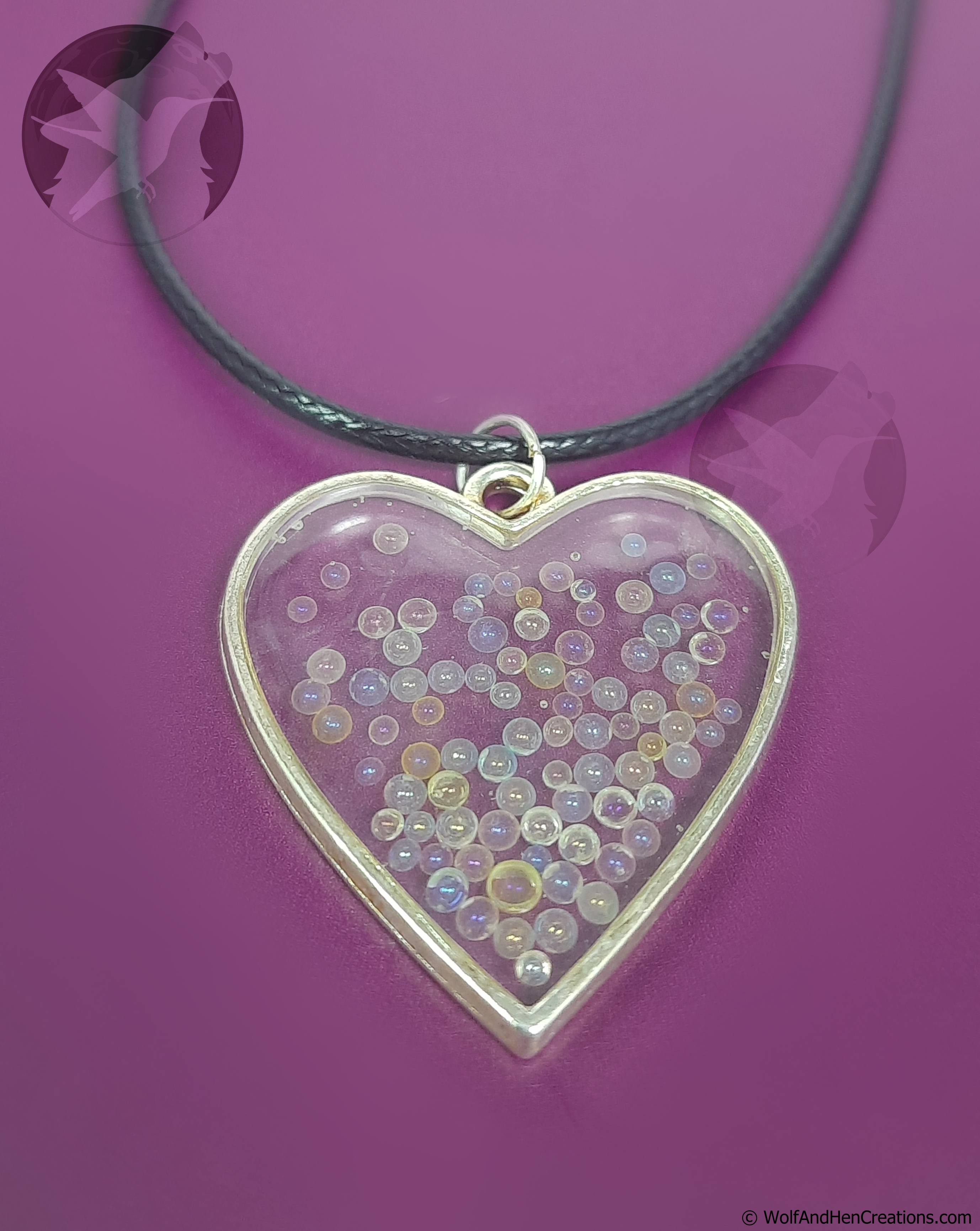 A heart shaped pendant necklace with a silver toned metal border and a clear centre filled with small, multicoloured iridescent glass beads, suspended from a black cord against a solid purple background.