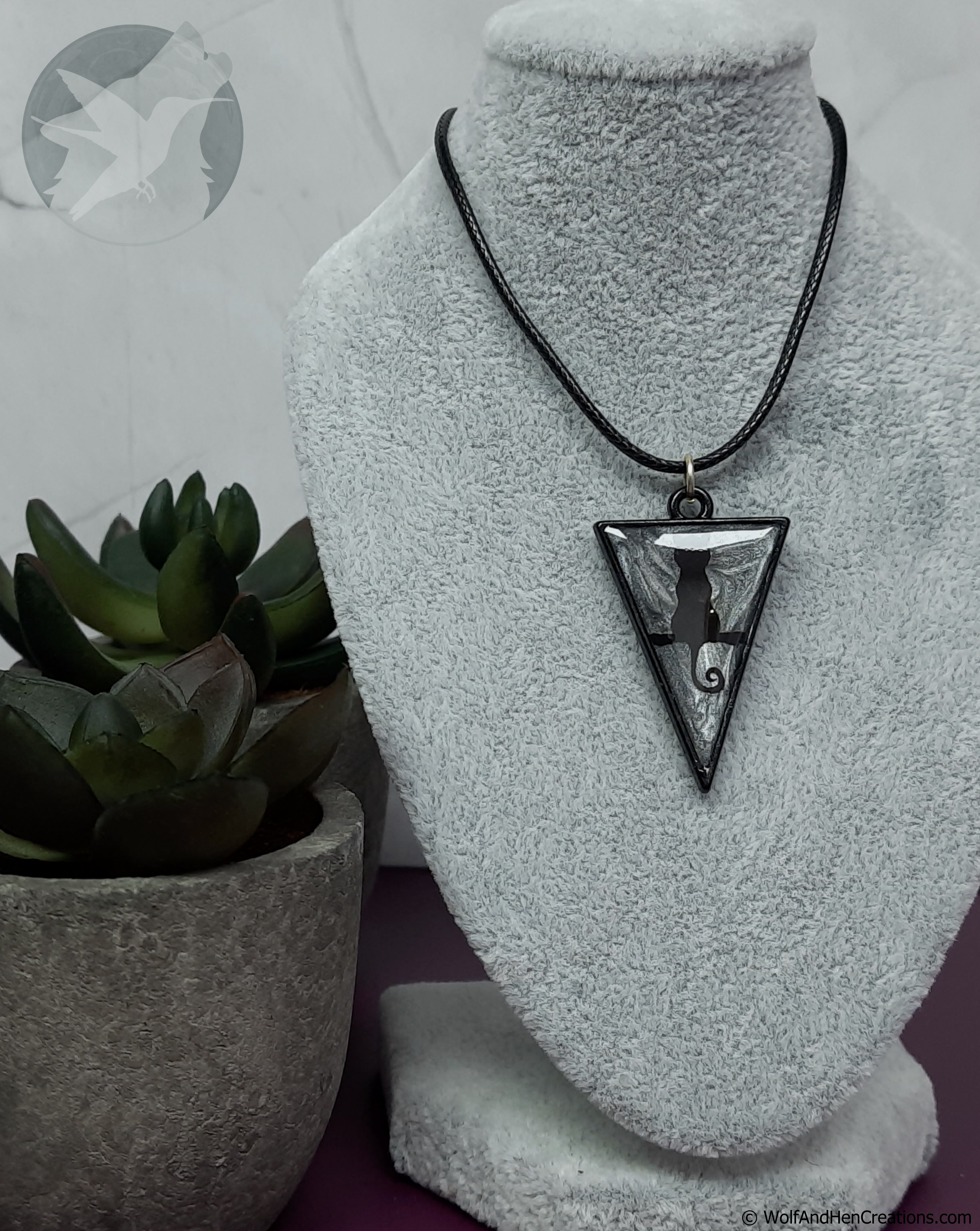A black and silver pendant necklace displayed on a grey velvet jewellery bust, featuring a triangular pendant with a black cat silhouette. A potted succulent sits to the left.