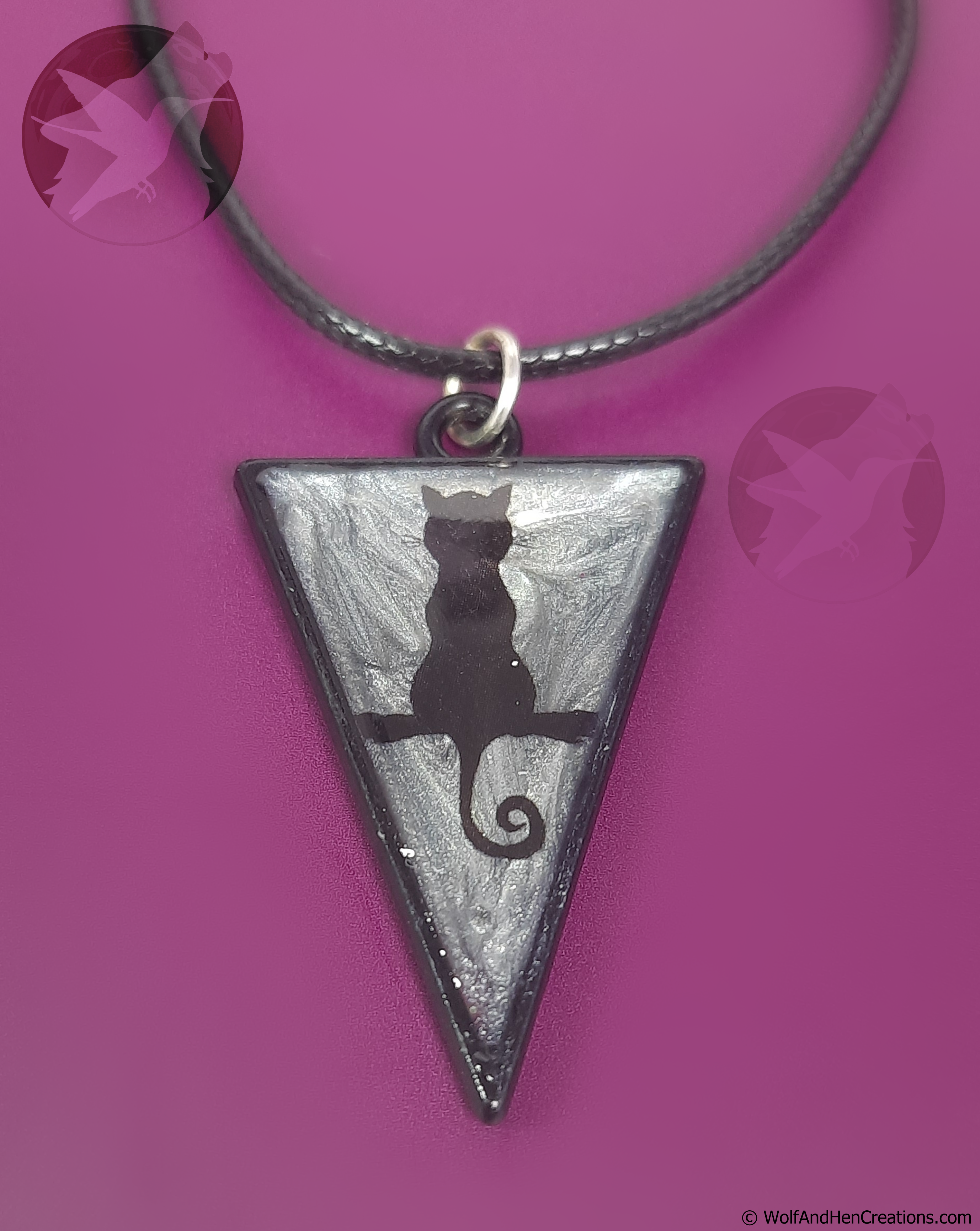 A triangular pendant necklace with a black cat silhouette on a silver background, hanging from a black cord against a bright purple surface.