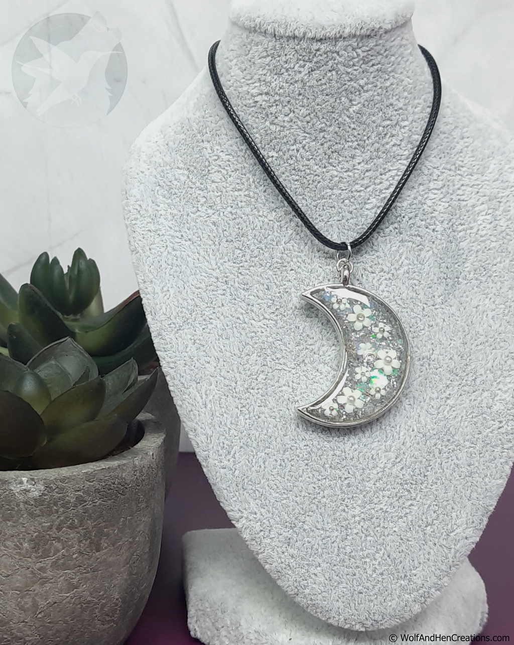 A silver crescent moon pendant necklace filled with iridescent glitter and white flowers, displayed on a grey textured jewellery bust. The necklace is suspended from a black cord.