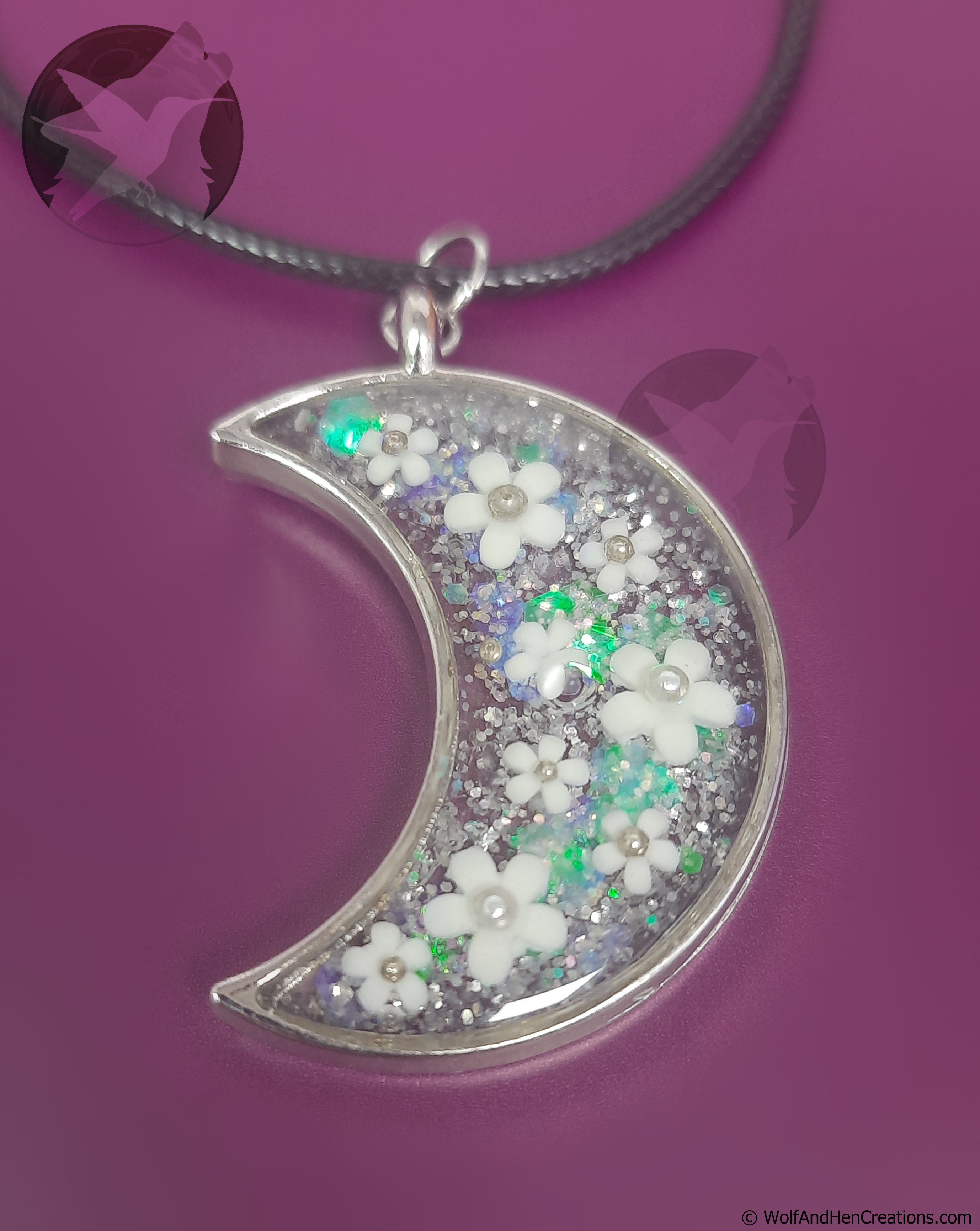 A crescent moon pendant necklace filled with white flower beads, iridescent glitter, and small faux pearl beads, hanging from a black cord against a solid purple background.