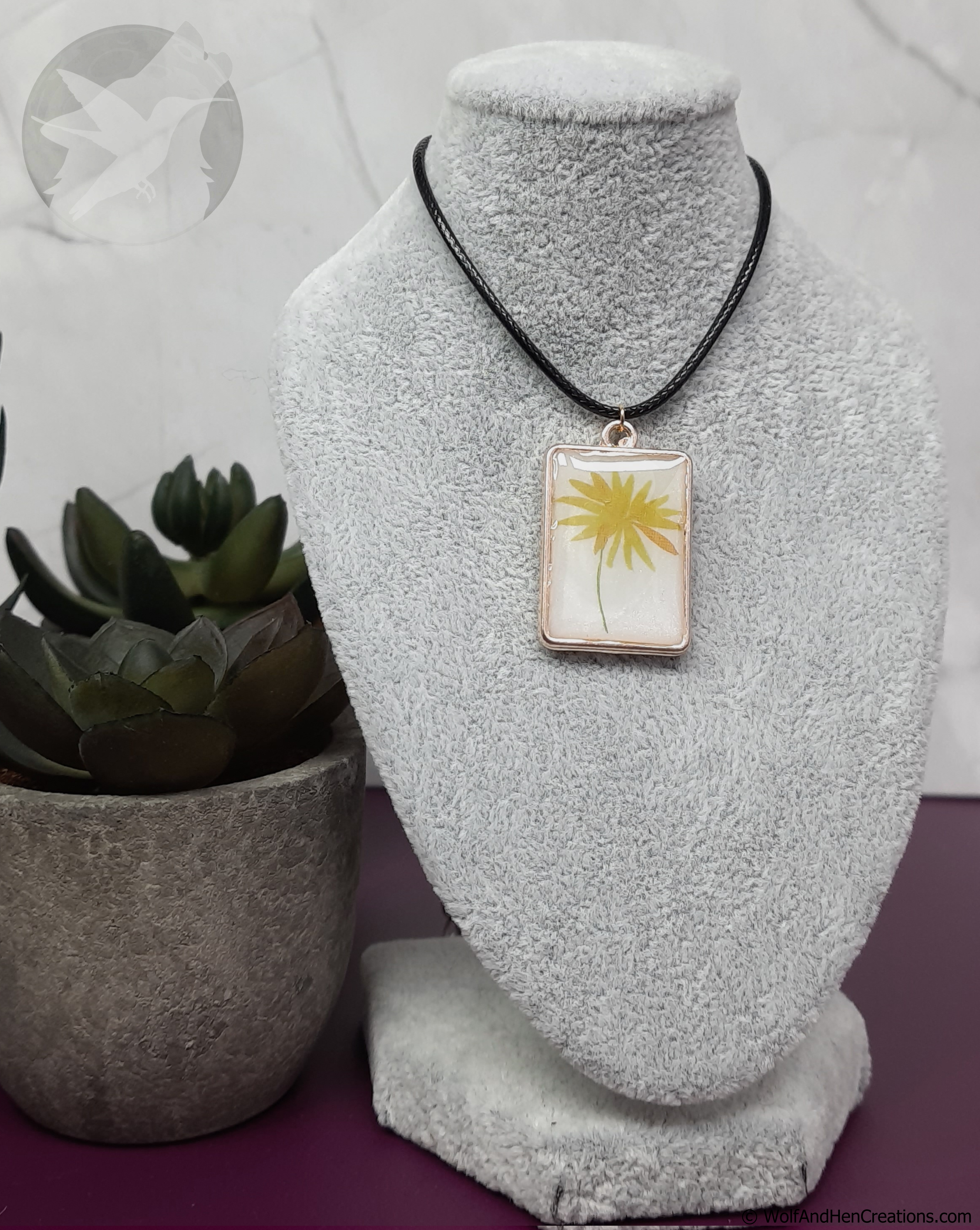 Necklace with a pendant featuring a yellow dandelion design on a textured stand with a plant in the background.