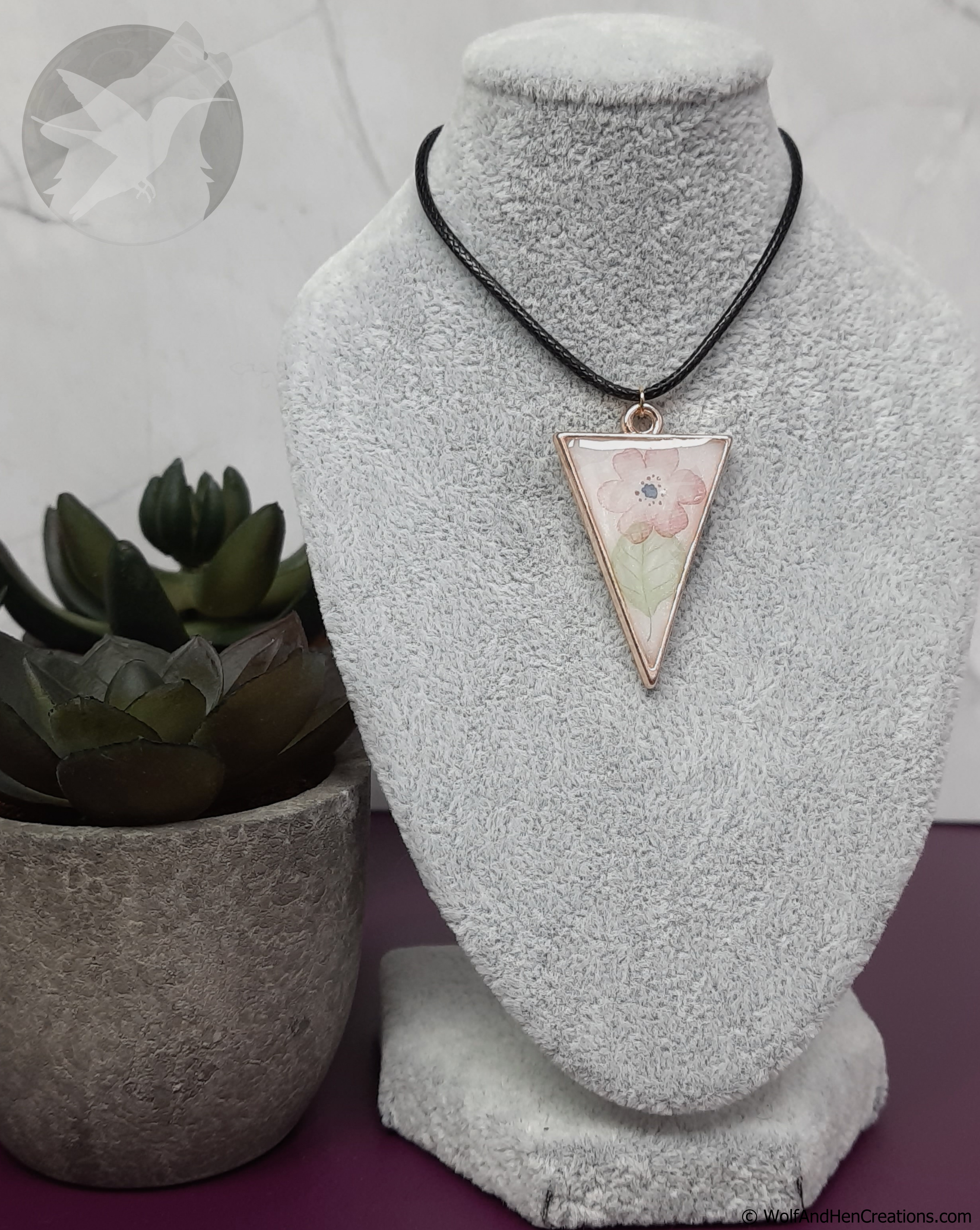 Necklace with a triangular pendant on a gray bust, next to a potted plant.