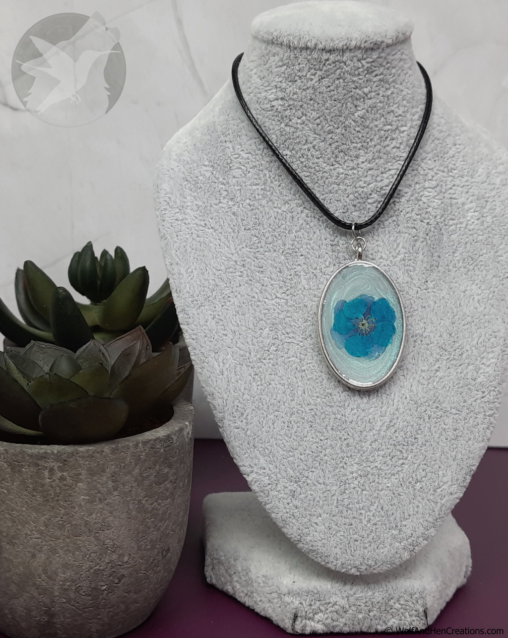 Necklace with a blue flower pendant on a white textured stand, next to a potted plant.