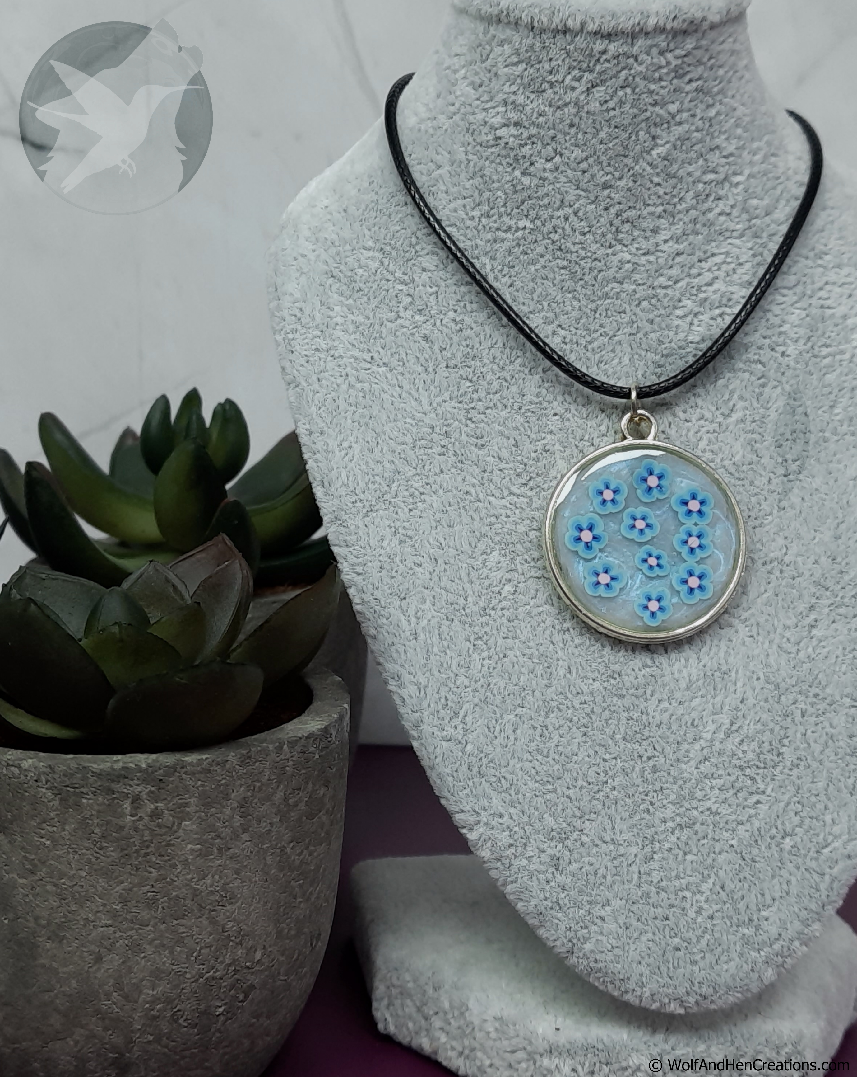 A round pendant necklace with a black cord and a silver-toned bezel, featuring light blue flowers with pink centres and a swirled iridescent blue-white background, displayed on a grey textured jewellery bust next to a potted succulent.