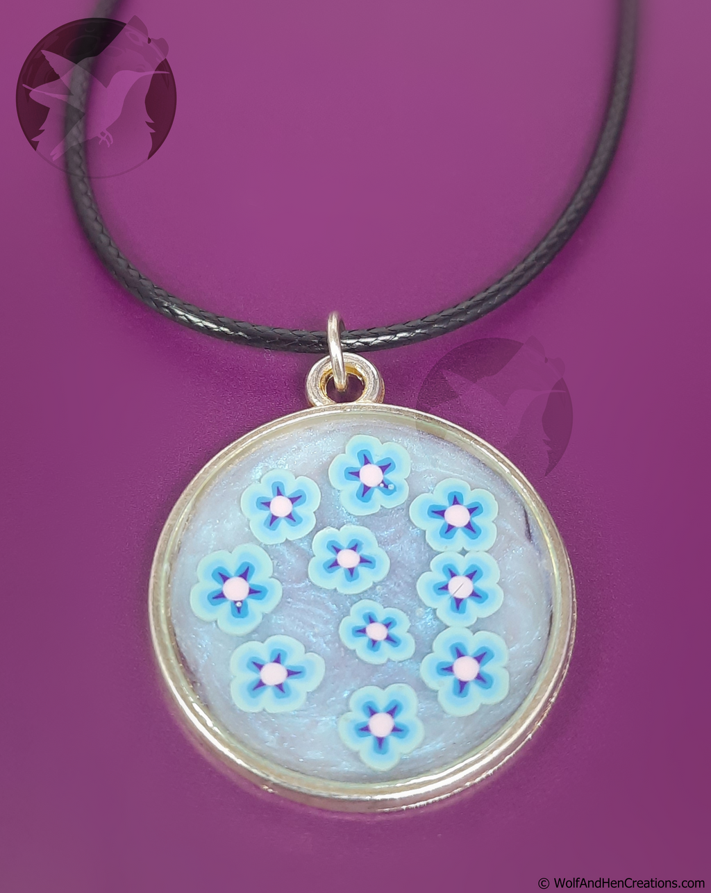A circular pendant necklace featuring light blue flowers with pink centres on a swirled iridescent blue-white base, suspended from a black cord on a solid purple background.