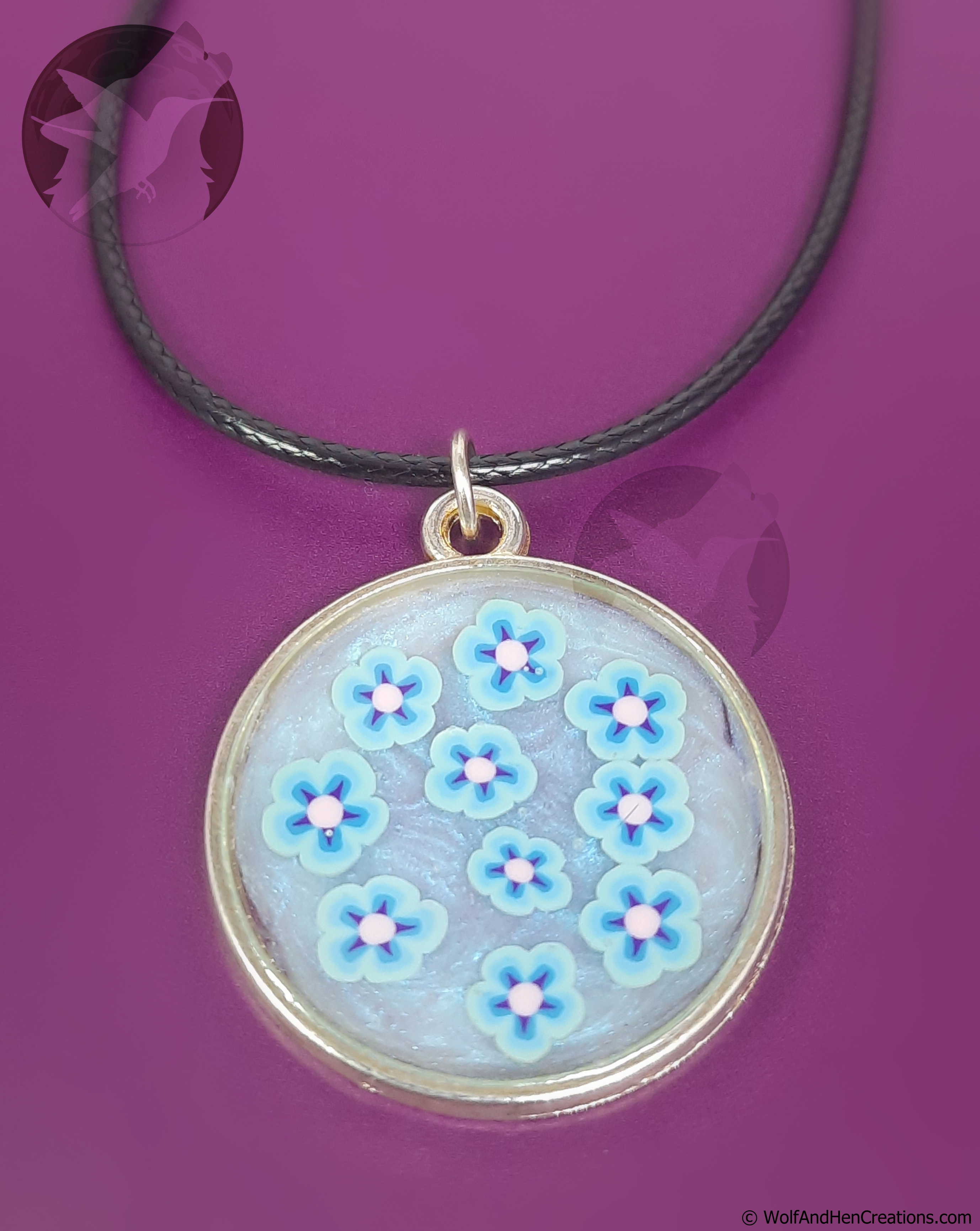 A circular pendant necklace featuring light blue flowers with pink centres on a swirled iridescent blue-white base, suspended from a black cord on a solid purple background.
