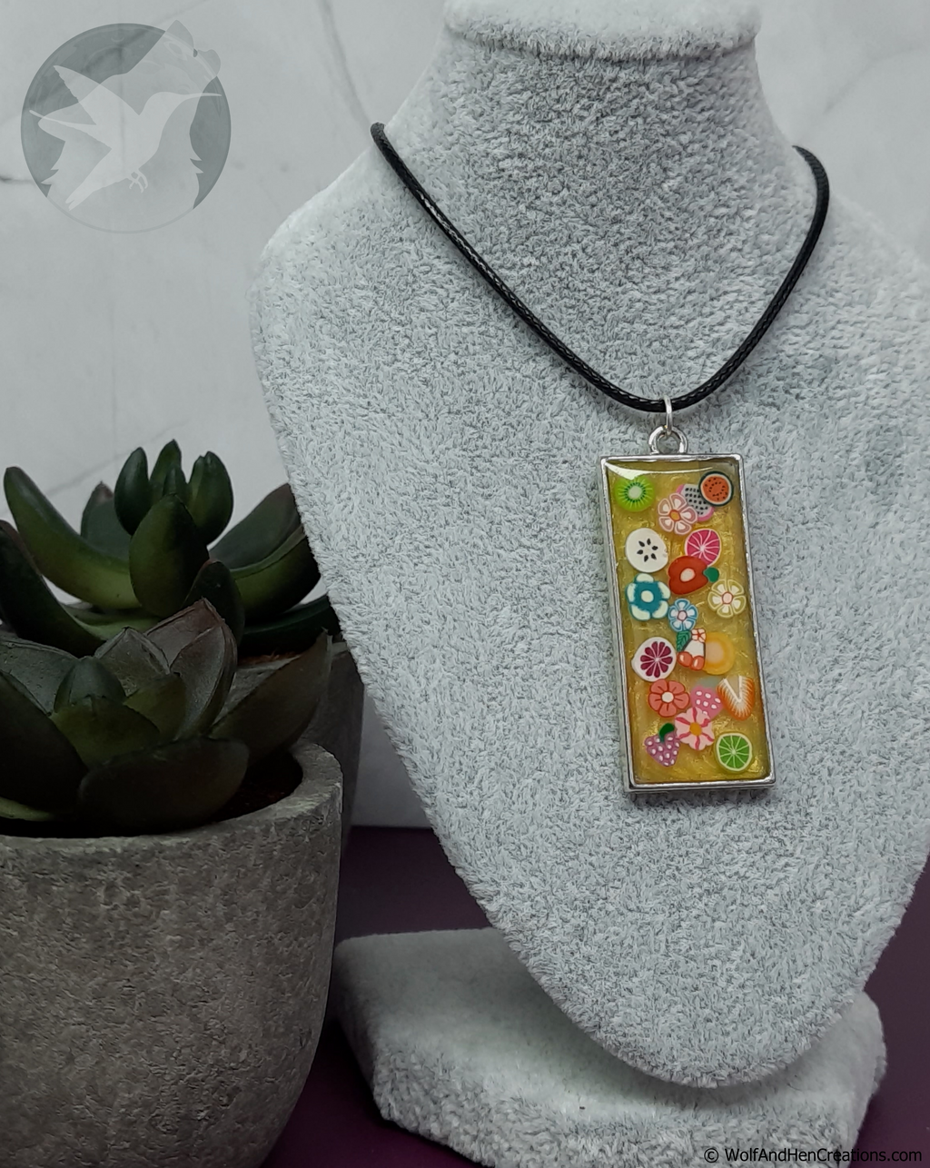 A rectangular pendant necklace with colourful floral and fruit slices on a yellow background, suspended from a black cord on a grey fabric jewellery bust. A small potted succulent is visible on the left.