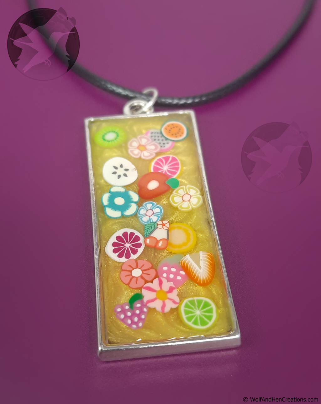 A rectangular pendant necklace with a silver frame, filled with a pearlescent yellow resin and decorated with colourful polymer clay flowers and fruits, hanging on a black cord against a solid purple background.