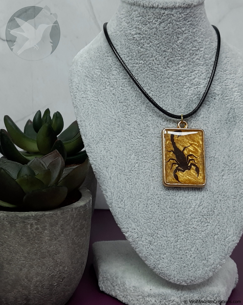 A rectangular pendant necklace displayed on a gray fabric jewelry bust. The pendant features a black scorpion design set against a textured gold background, suspended from a black cord.