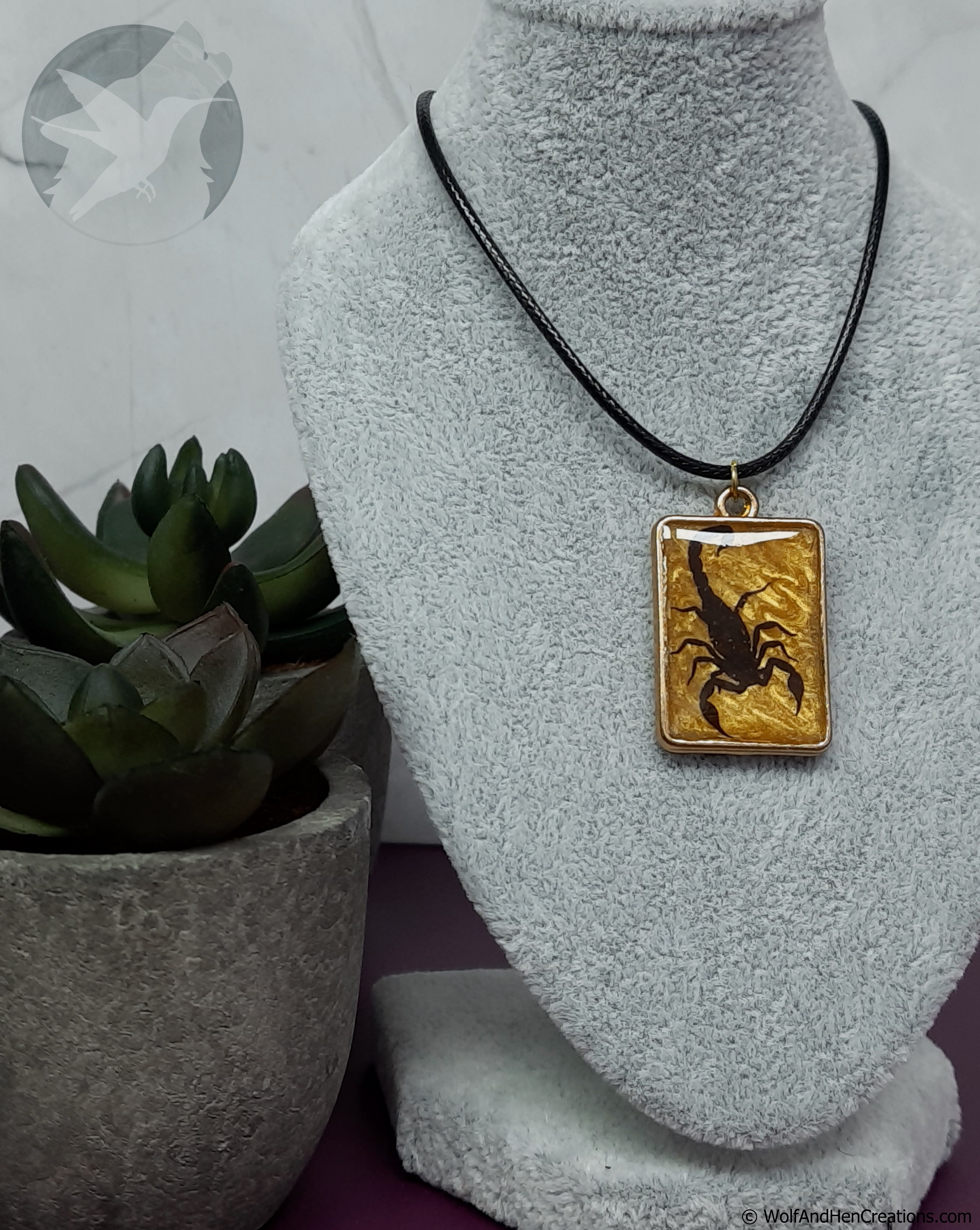 A rectangular pendant necklace displayed on a gray fabric jewelry bust. The pendant features a black scorpion design set against a textured gold background, suspended from a black cord.