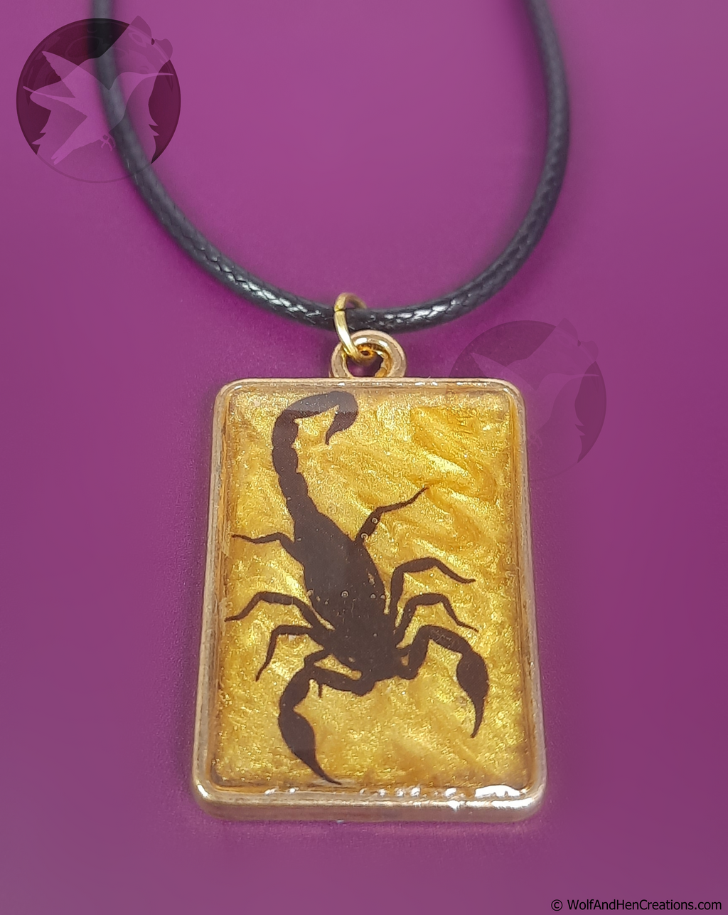 A rectangular pendant necklace featuring a black scorpion silhouette over a textured gold background, suspended from a black cord, set against a solid purple background.
