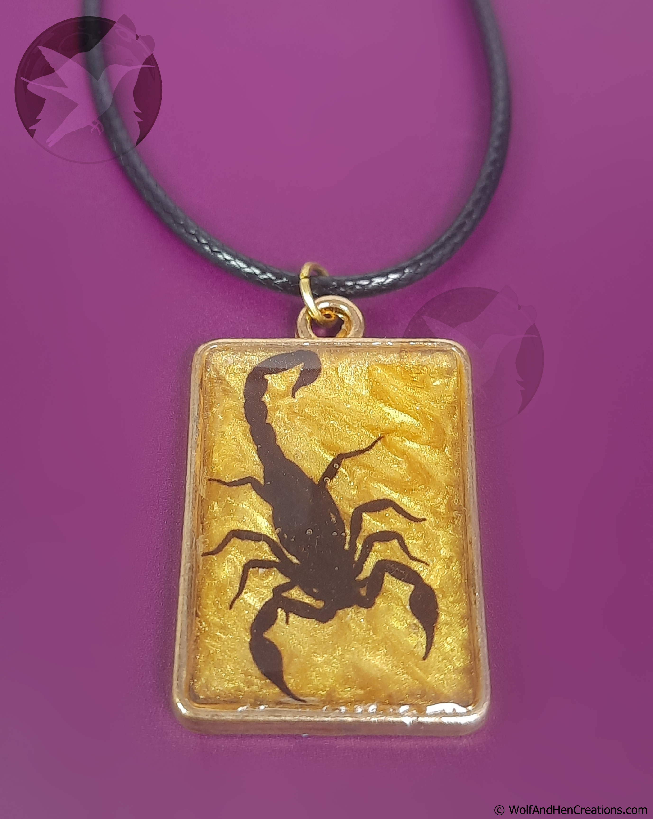 A rectangular pendant necklace featuring a black scorpion silhouette over a textured gold background, suspended from a black cord, set against a solid purple background.
