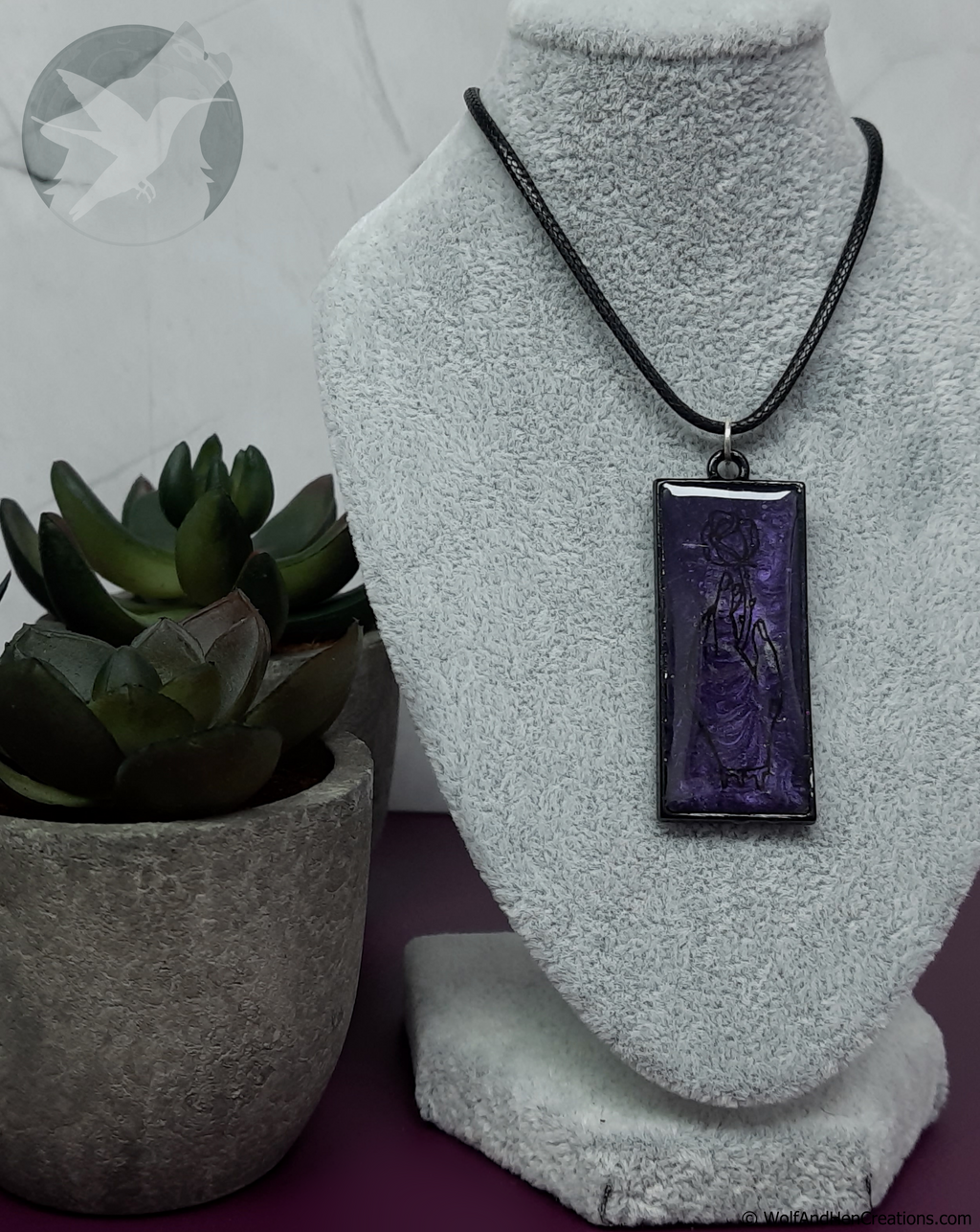 A rectangular purple pendant necklace with a black hand grasping a cut rose, displayed on a grey textured jewellery bust. The necklace has a black cord and is styled next to a small potted succulent.