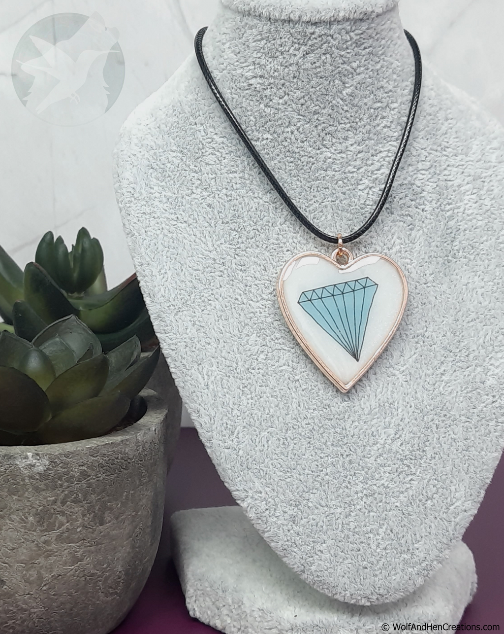 A heart shaped pendant necklace displayed on a grey fabric mannequin bust. The pendant has a subtly shimmering white resin background with a light blue diamond design and a rose gold border, hanging from a black cord.
