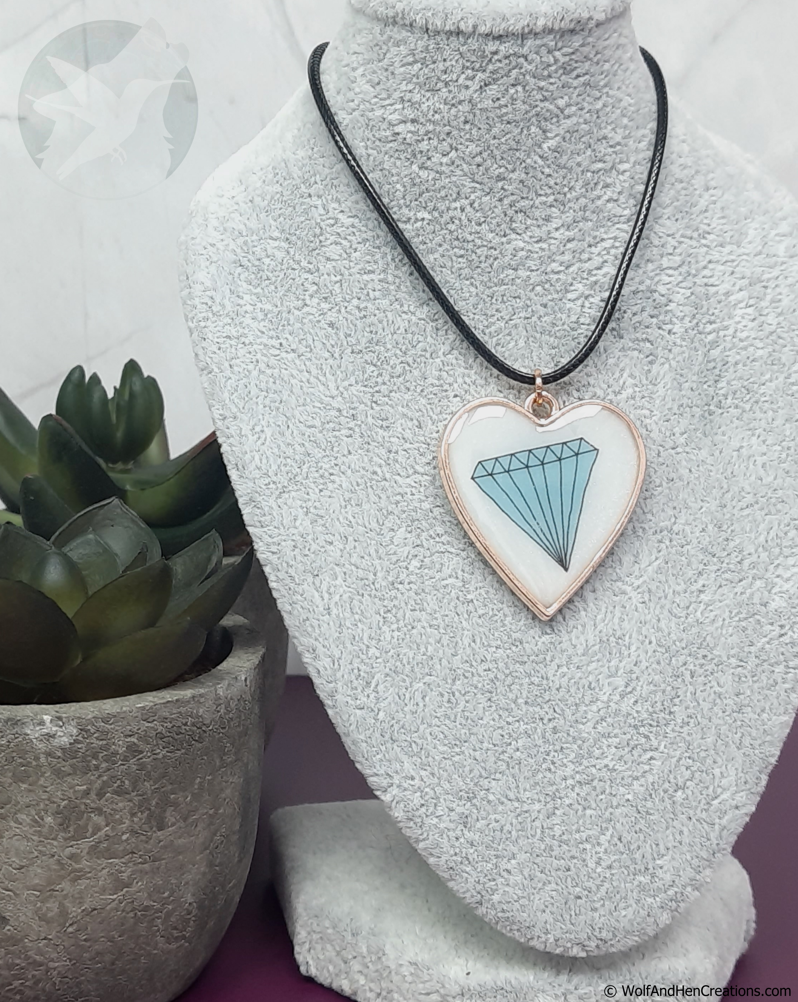 A heart shaped pendant necklace displayed on a grey fabric mannequin bust. The pendant has a subtly shimmering white resin background with a light blue diamond design and a rose gold border, hanging from a black cord.