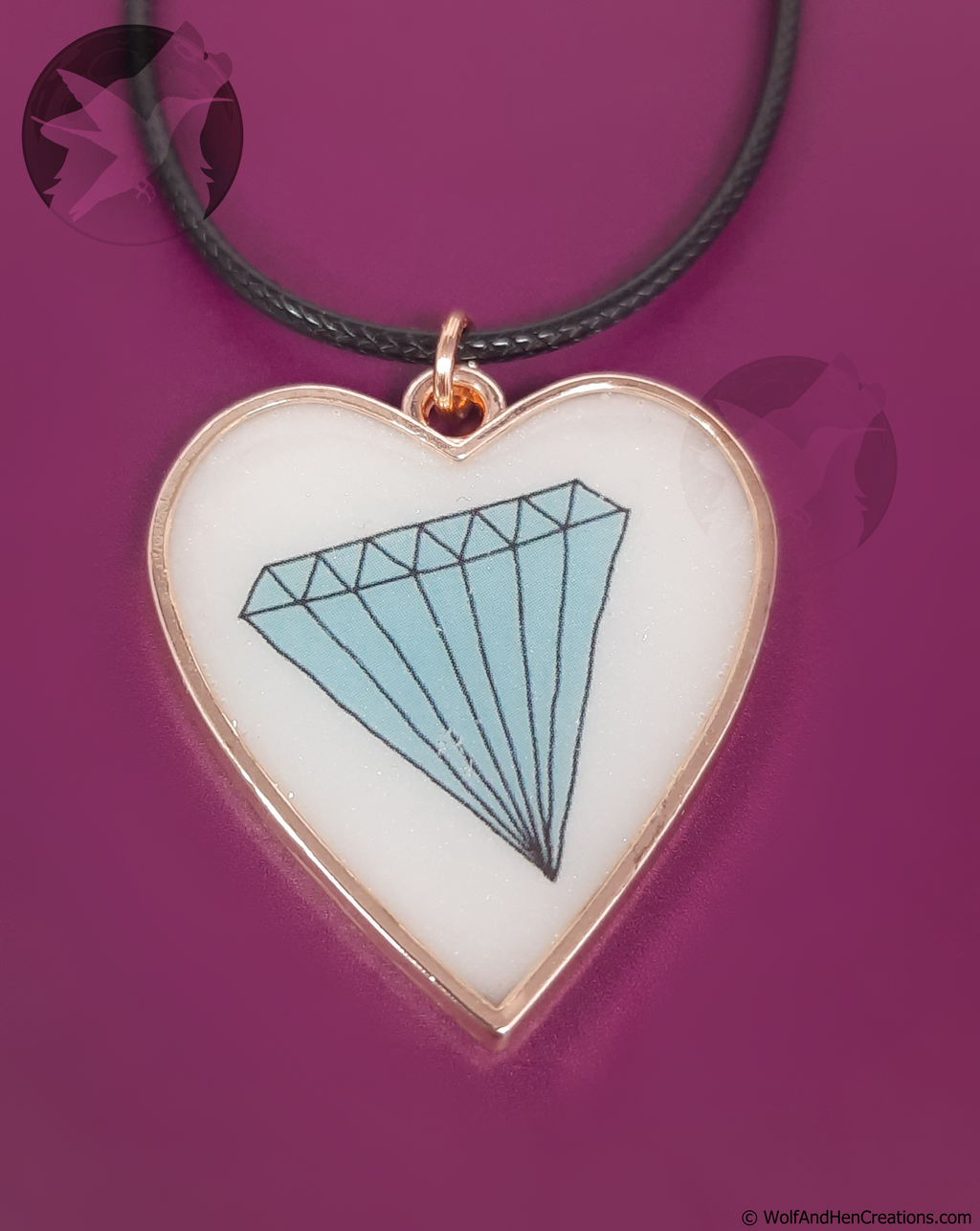 A heart shaped pendant necklace featuring a subtly iridescent white background with a light blue diamond illustration and a rose gold border, suspended from a black cord against a magenta background.