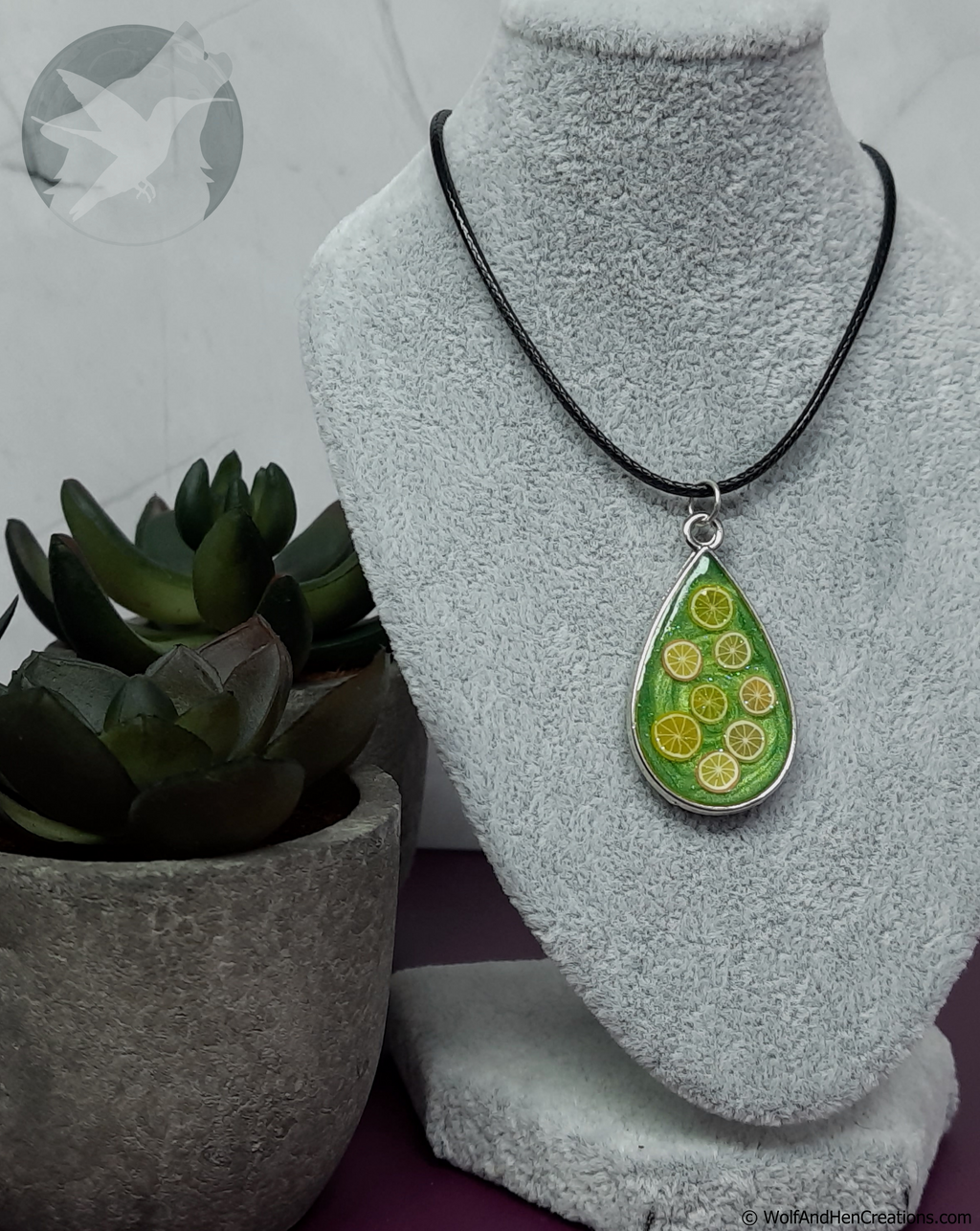 A teardrop-shaped pendant necklace with a green citrus design hangs on a black cord from a gray display bust, with a potted succulent in the background.