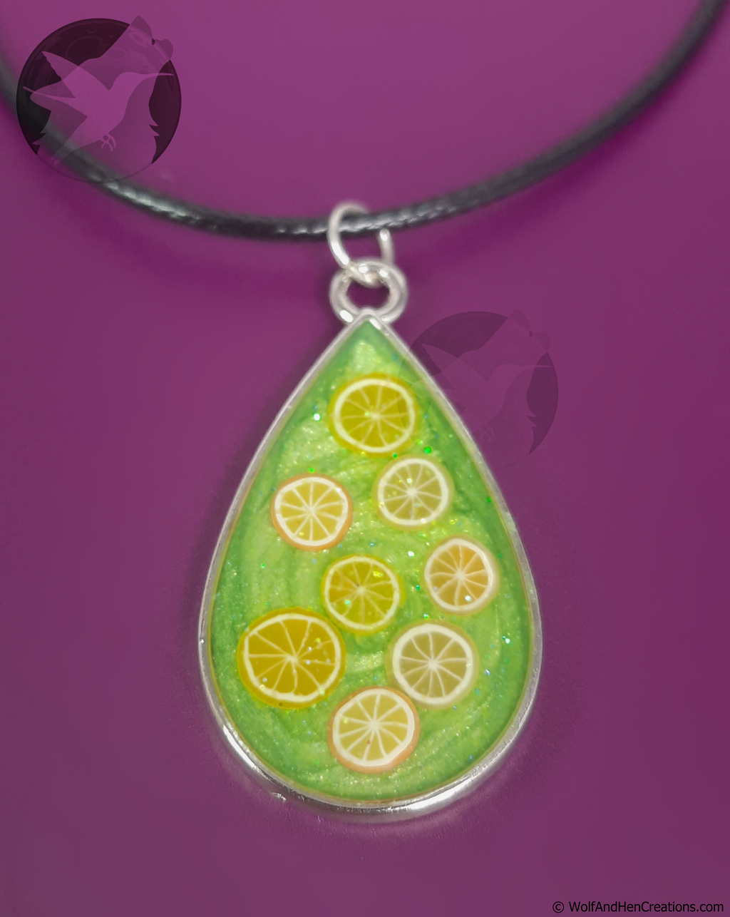 A teardrop-shaped pendant necklace featuring a green glittery background with lemon and orange clay slices, suspended from a black cord against a solid purple background.