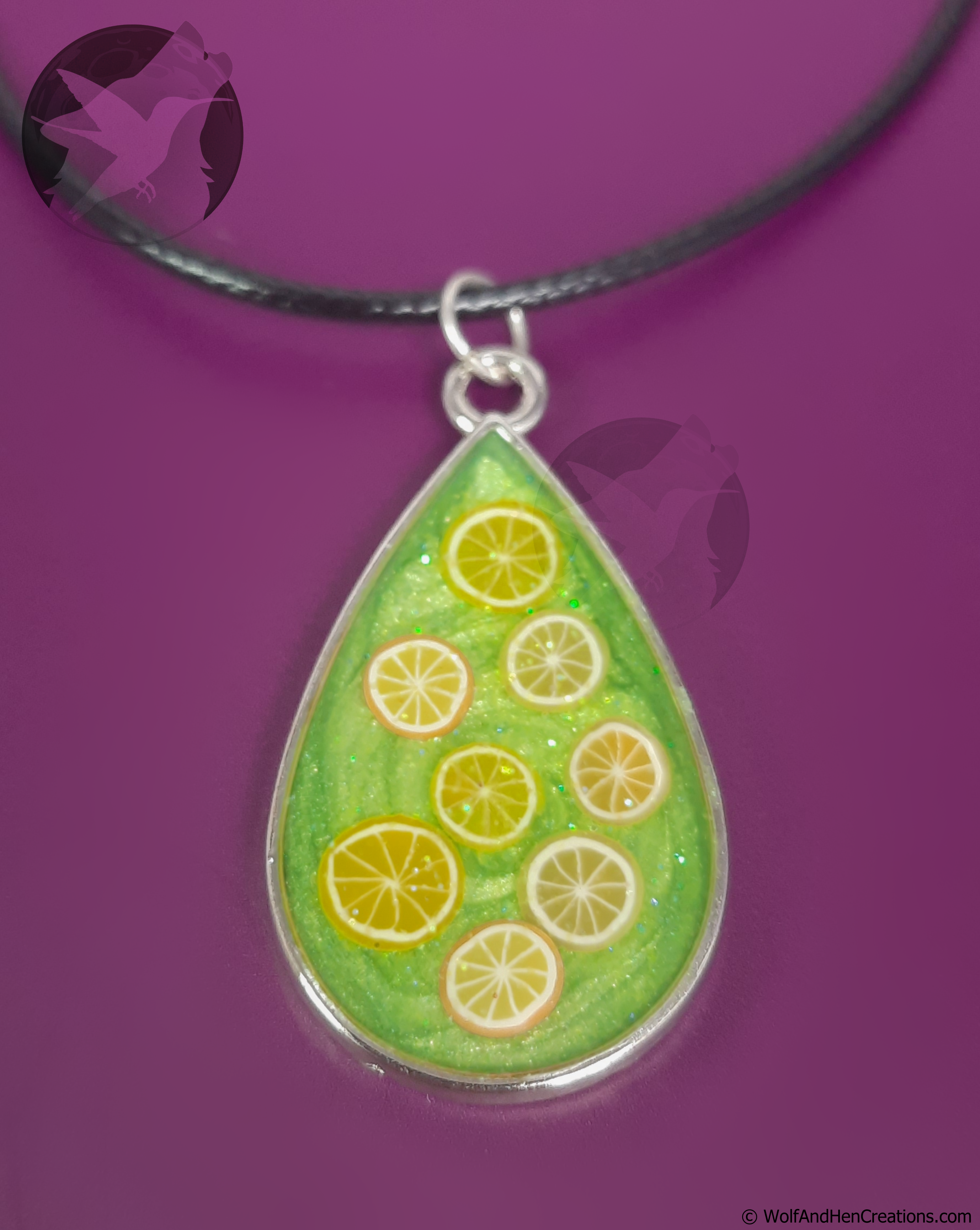 A teardrop-shaped pendant necklace featuring a green glittery background with lemon and orange clay slices, suspended from a black cord against a solid purple background.