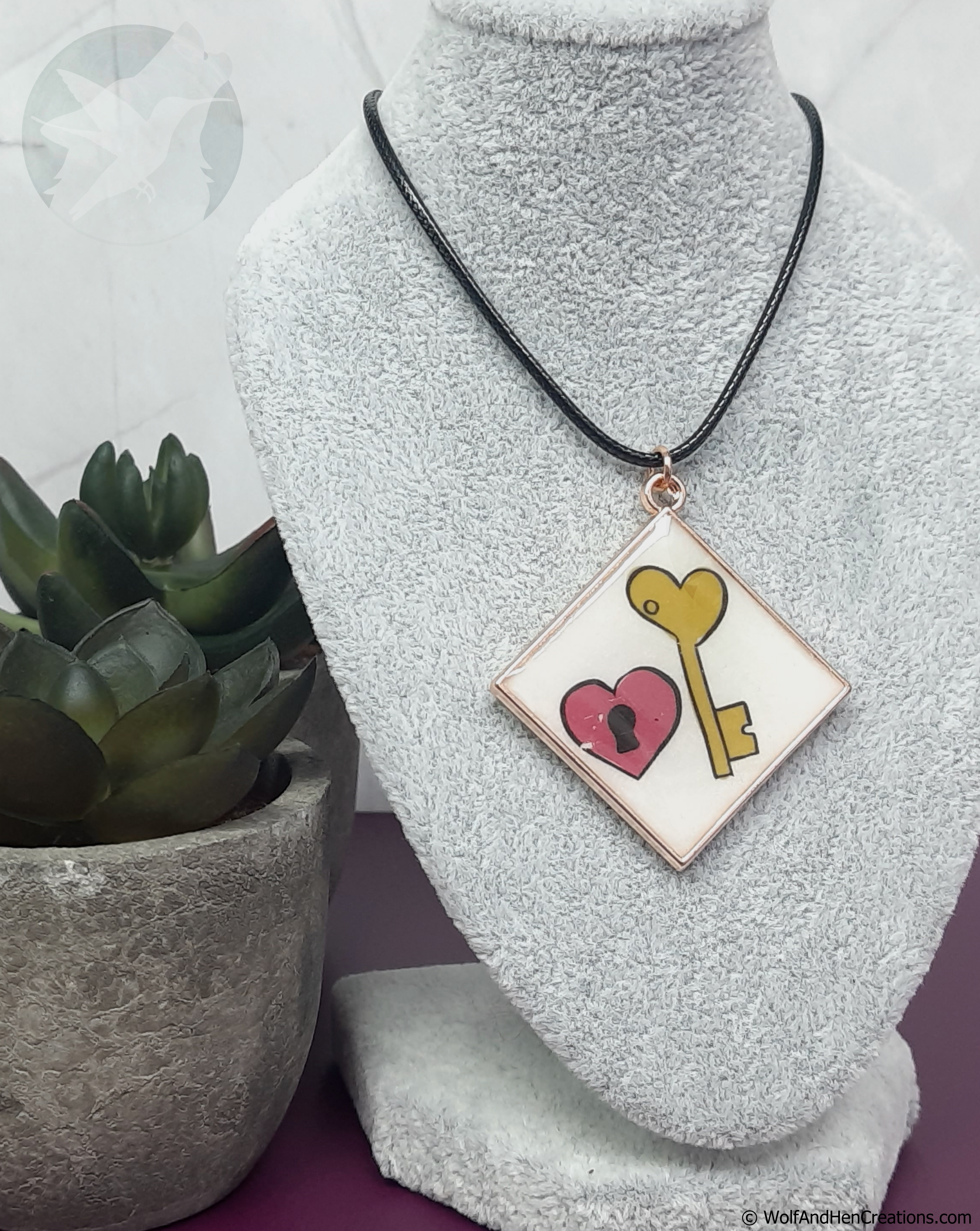 A square shaped pendant necklace displayed on a grey textured mannequin bust. The pendant has a white background with a yellow heart shaped key and a pink heart with a black lock, bordered in gold metal and suspended from a black cord.