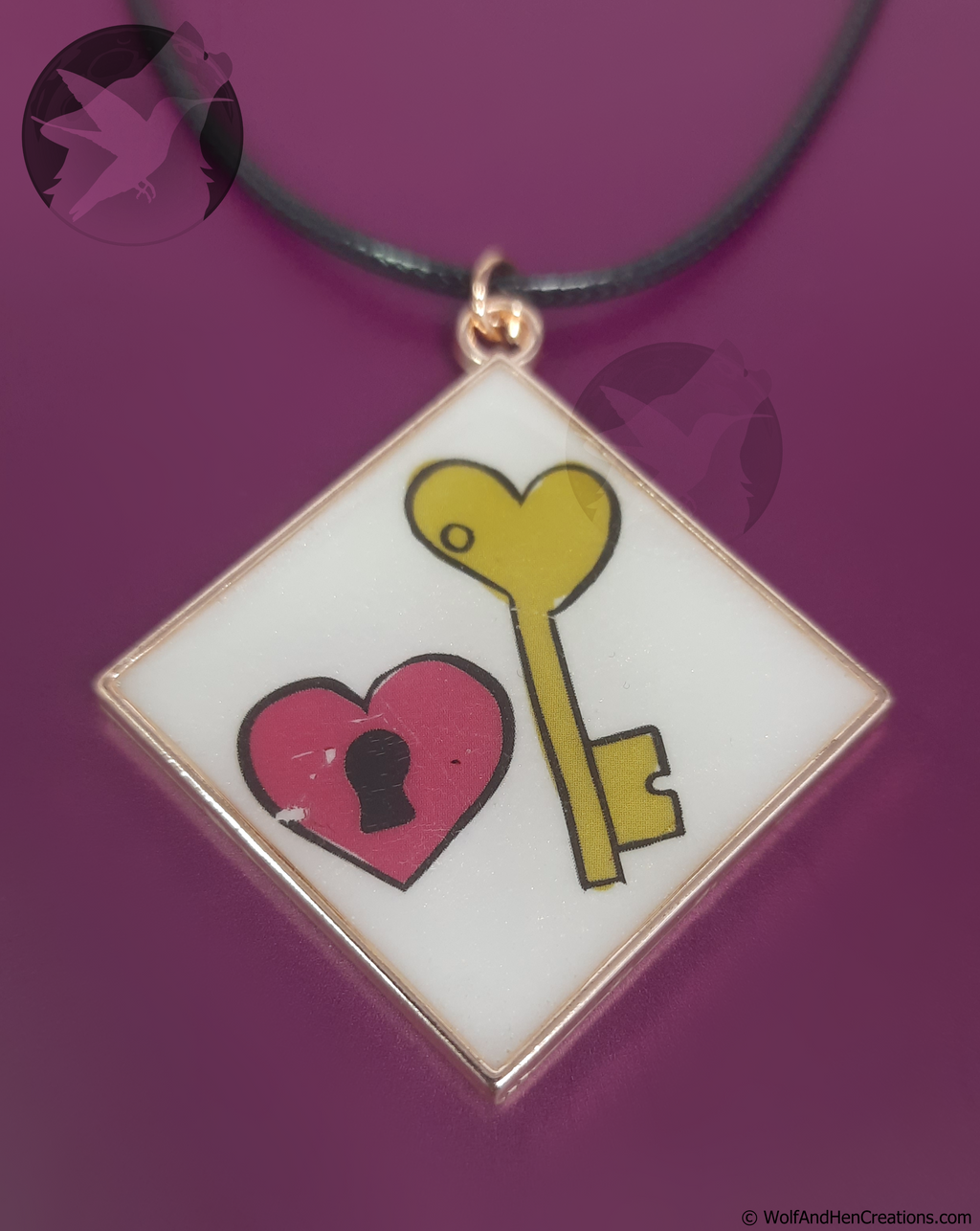 A square pendant with an iridescent white face, featuring a yellow heart shaped key and a pink heart with a black keyhole designs, suspended on a black cord against a solid purple background.