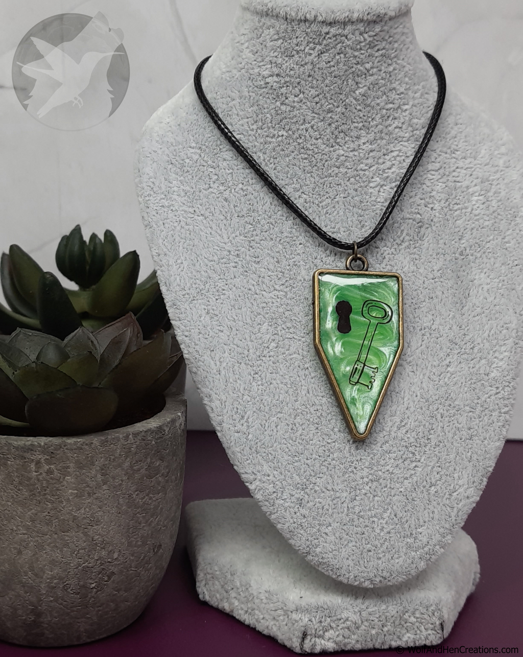 Necklace with a green pendant featuring a key design on a gray stand with a plant in the background.