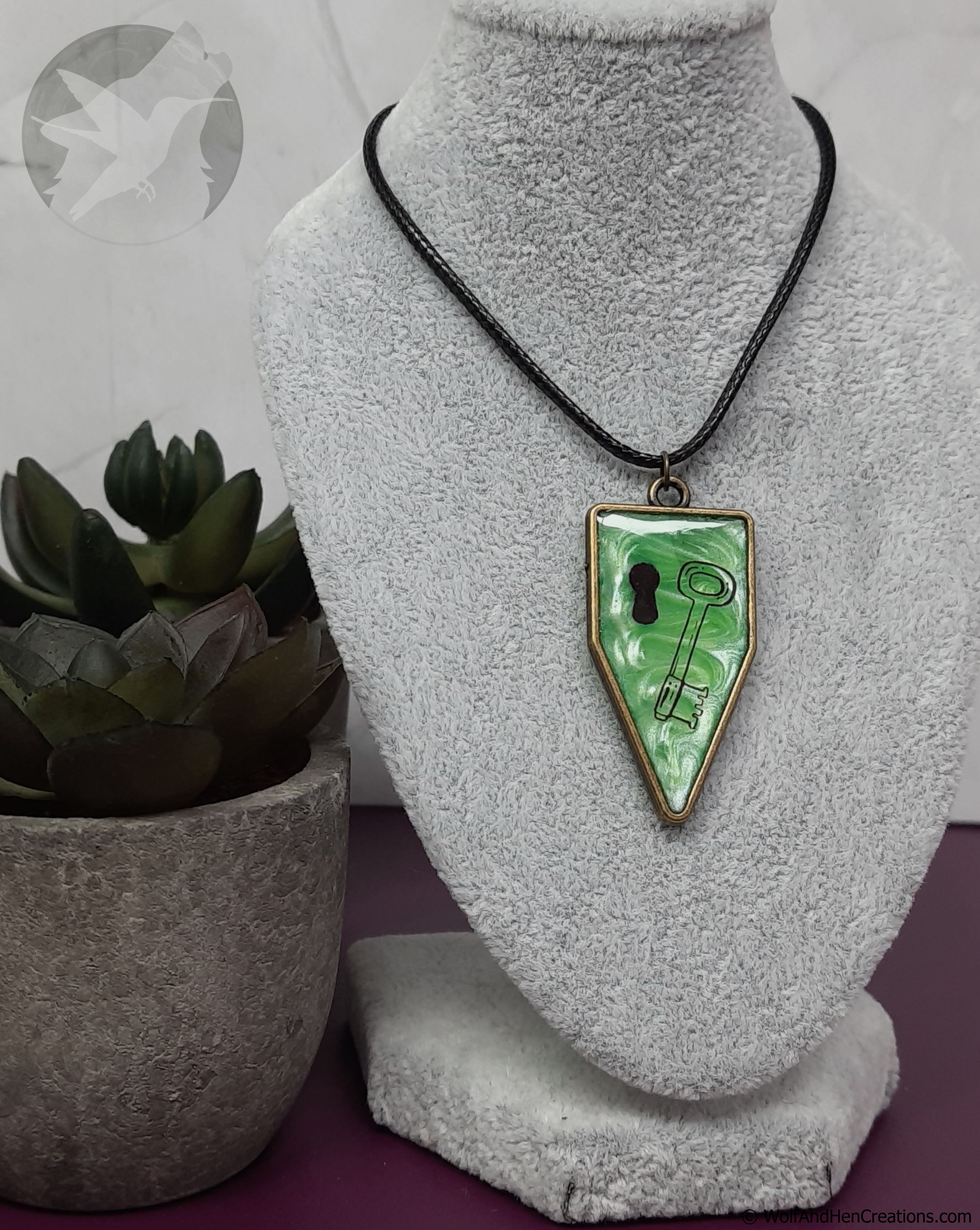 Necklace with a green pendant featuring a key design on a gray stand with a plant in the background.