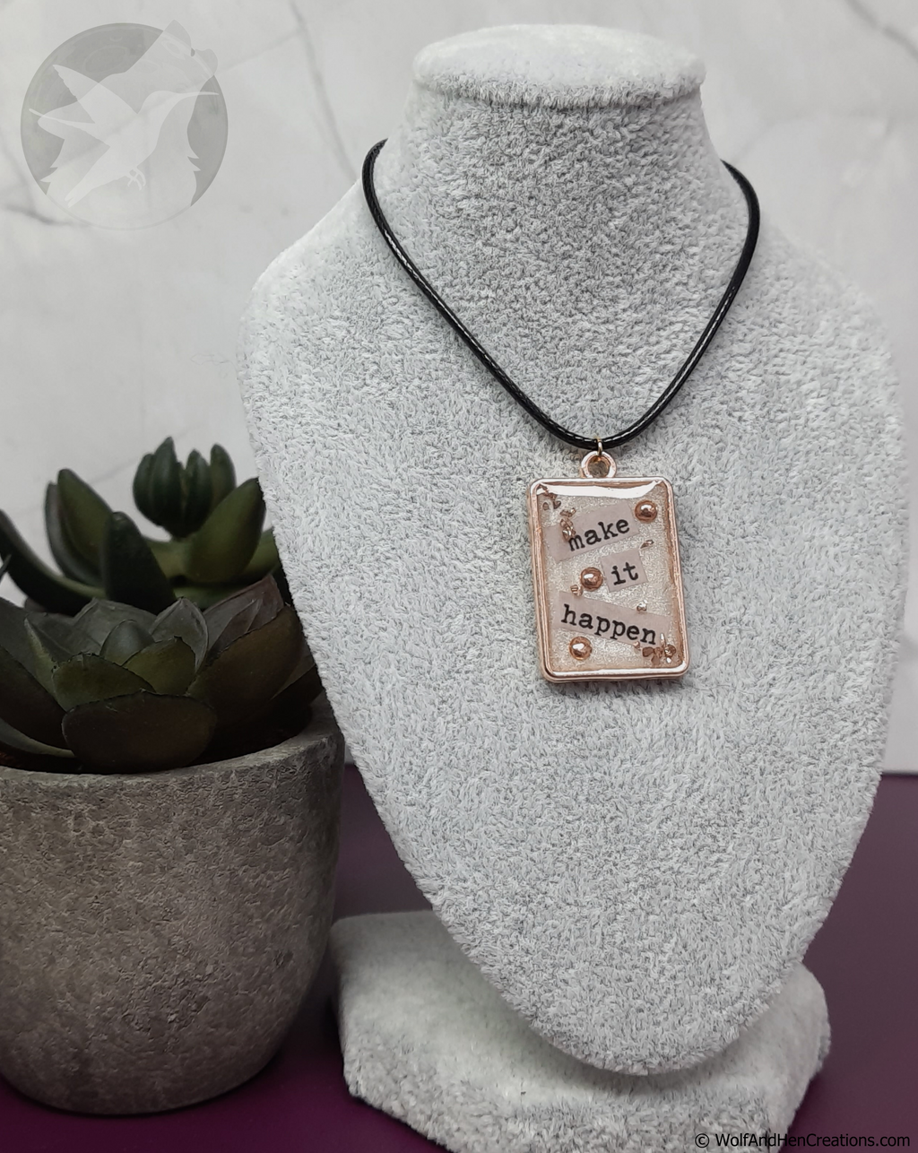 Necklace with motivational pendant that says "make it happen" on a white bust, next to a potted plant.