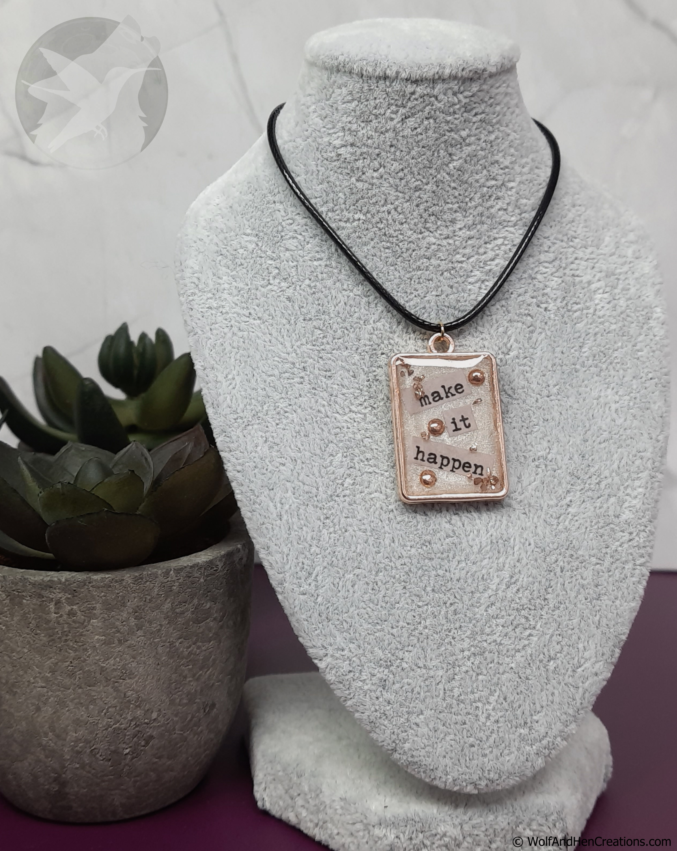 Necklace with motivational pendant that says "make it happen" on a white bust, next to a potted plant.