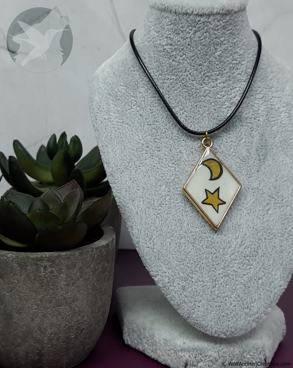 A necklace with a black cord and a diamond-shaped pendant with a white background, gold border with a yellow crescent moon and star. A potted succulent is in the background.