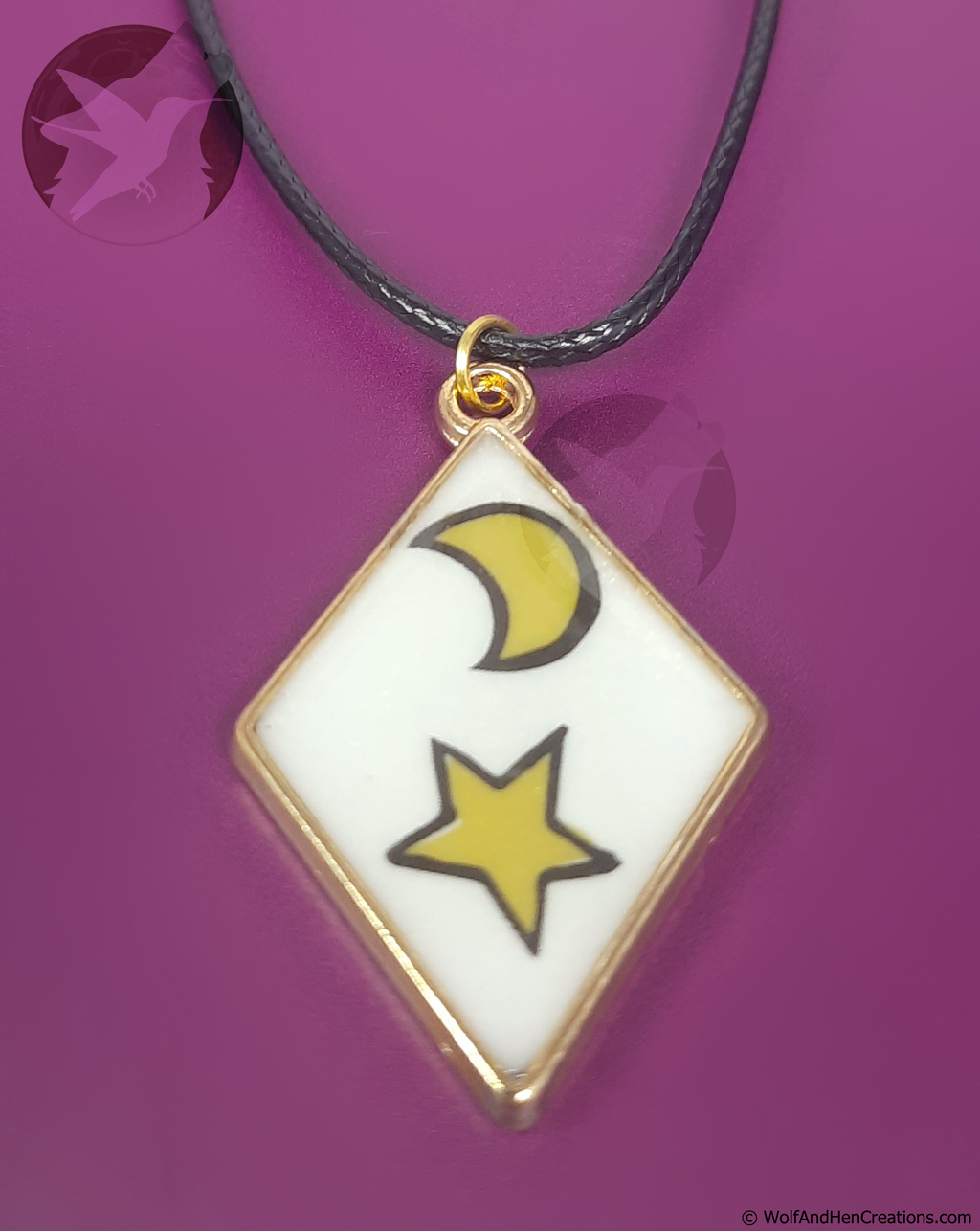 A diamond-shaped pendant necklace featuring a white background with a yellow crescent moon and star outlined in black, suspended from a black cord with a gold-tone ring all on a solid magenta-purple background.