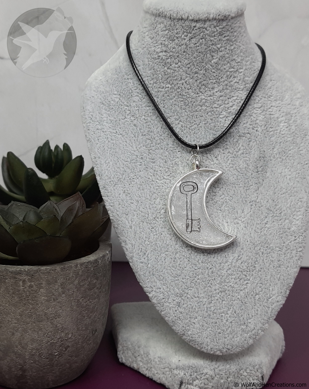 Silver crescent moon key pendant on a black cord displayed on a gray bust with a plant in the background.