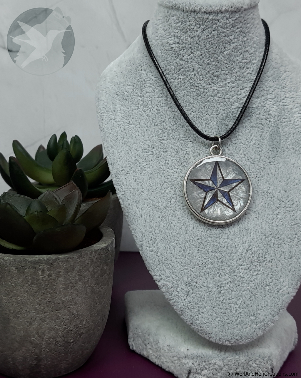 A silver circular pendant necklace featuring a blue and black five-pointed star design, displayed on a grey fabric jewellery bust with a small potted succulent in the background.